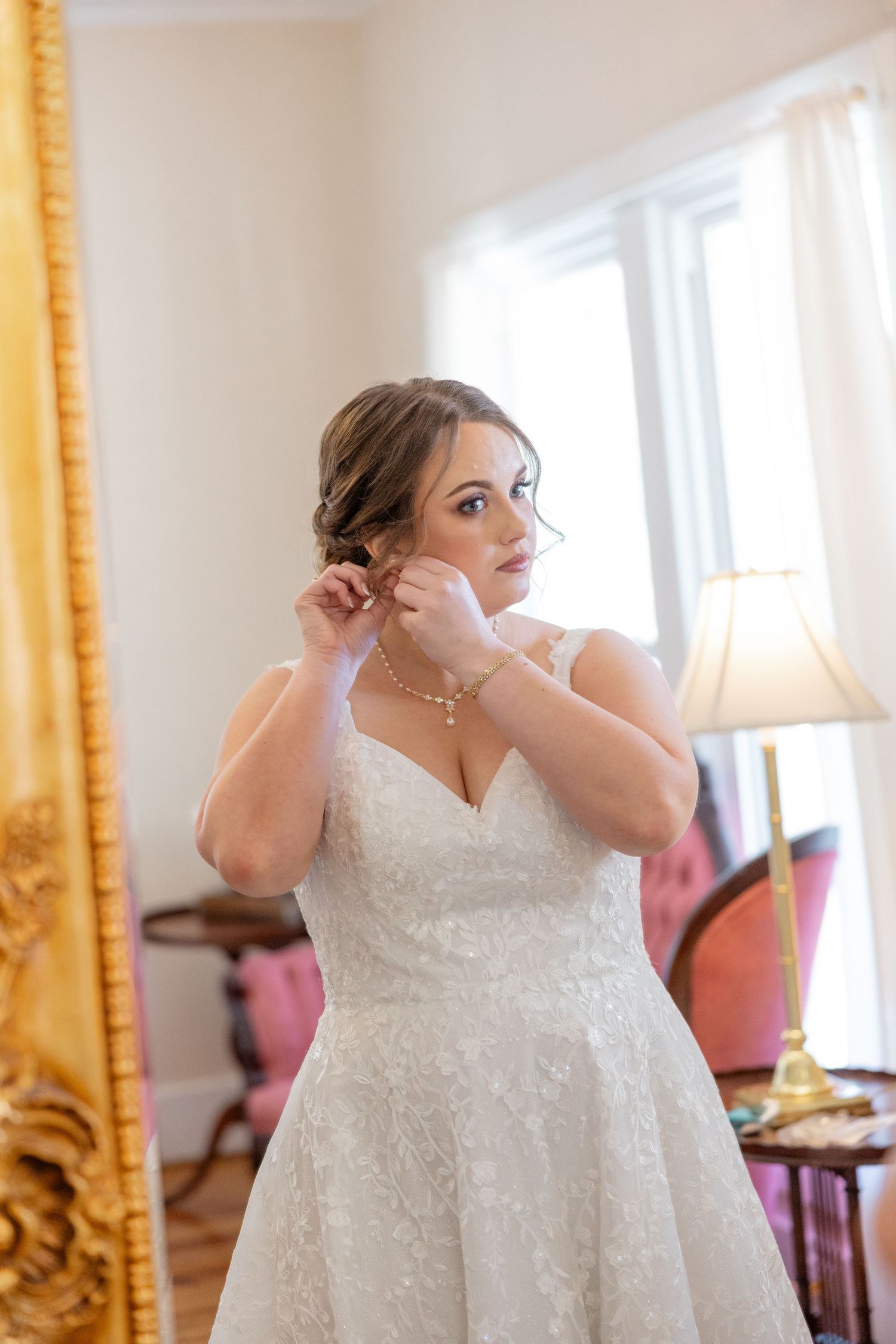 A white lace wedding dress displayed in a bright room with ornate gold-framed mirrors.