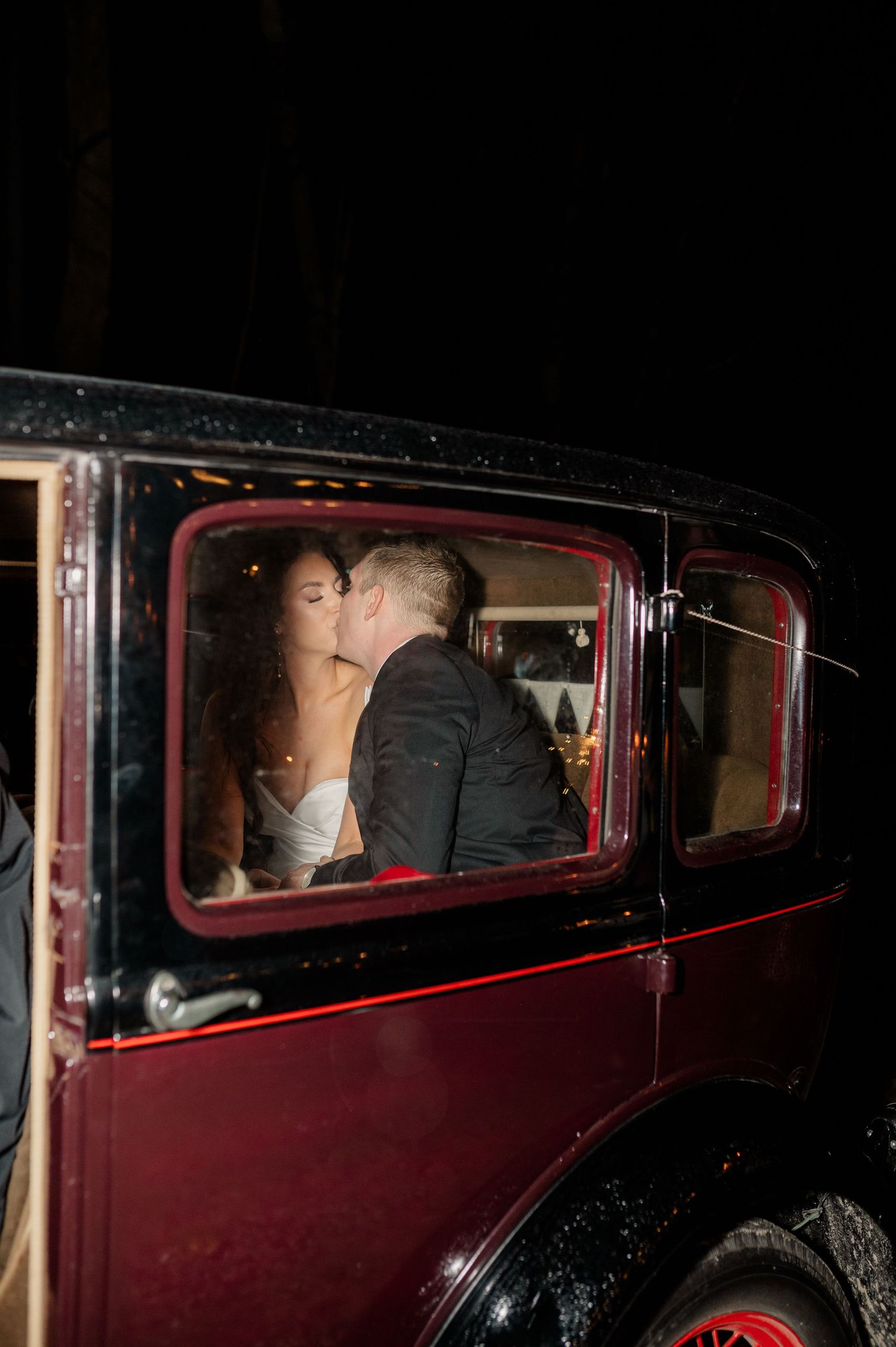 A vintage burgundy car serves as the wedding getaway vehicle in a nighttime setting.