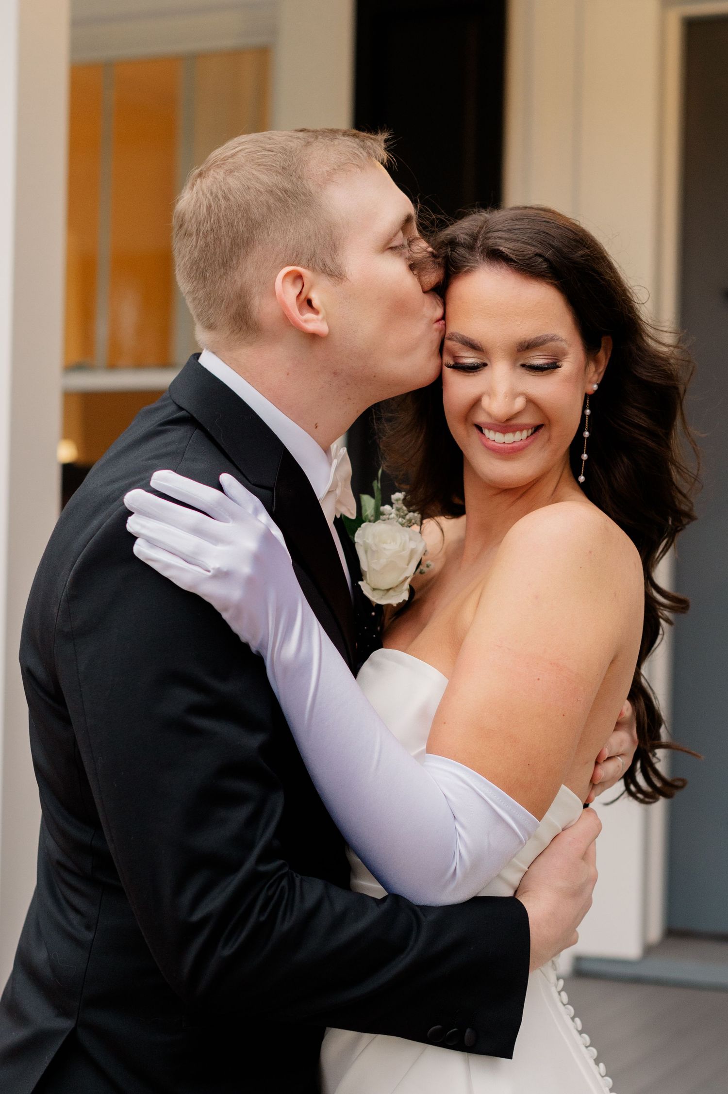 A couple shares intimate moments in formal wedding attire against a white building backdrop.