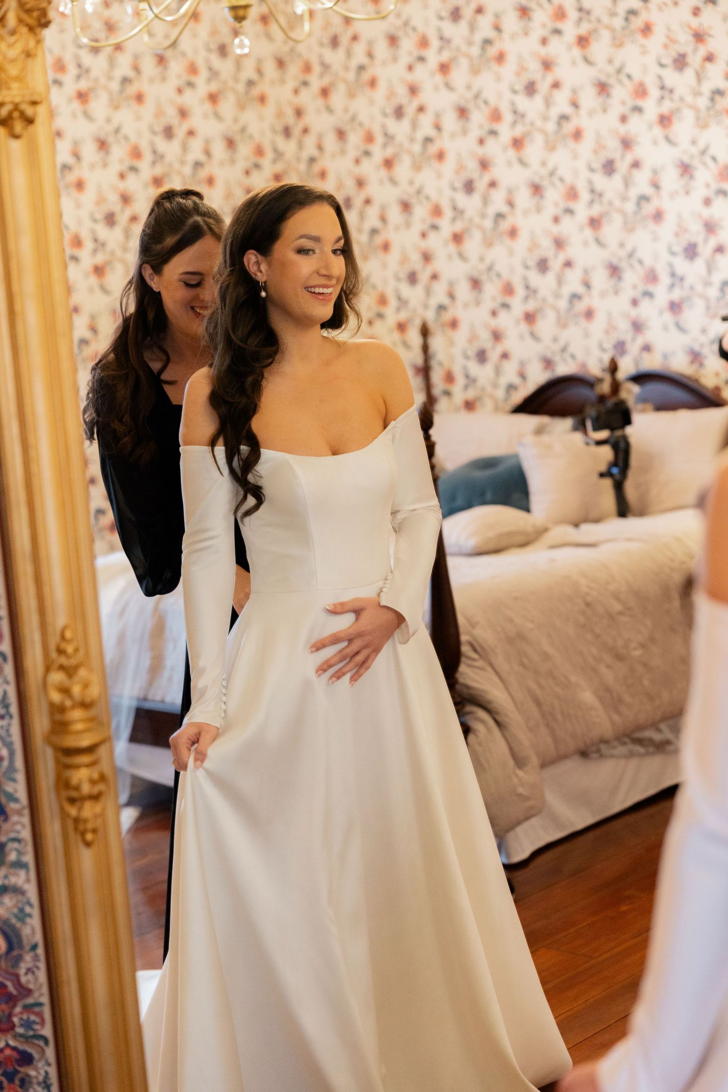 Bride in a classic white off-the-shoulder wedding gown twirls joyfully in a room with floral wallpaper.
