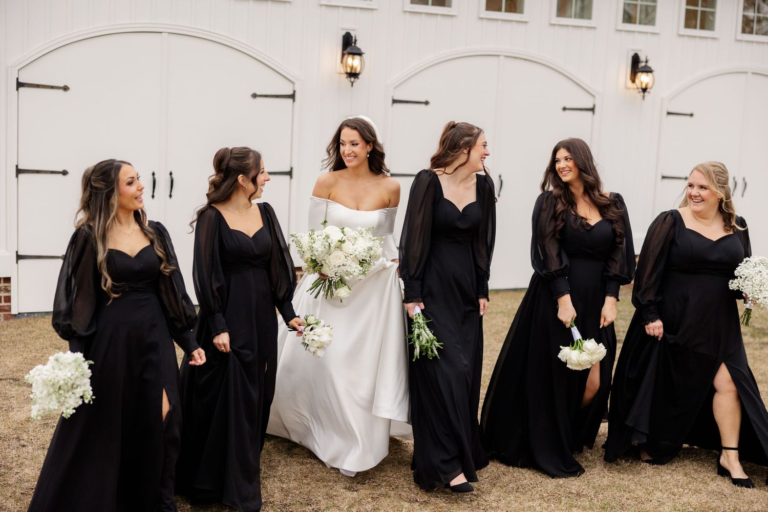 Wedding party in coordinated black dresses and white gown pose against white building exterior.