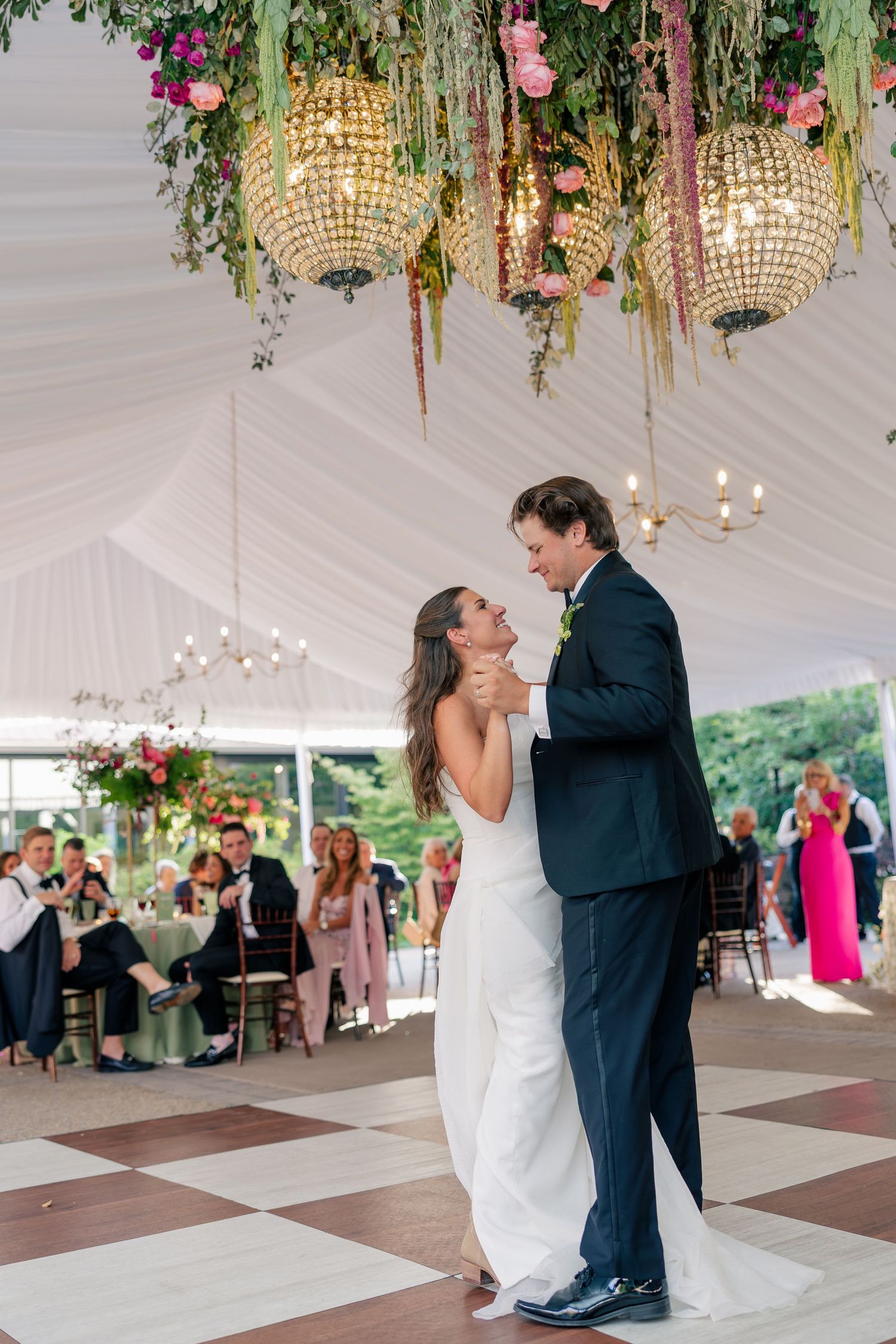 Bride and groom share a joyful first dance beneath lush floral chandeliers in an elegant wedding tent.