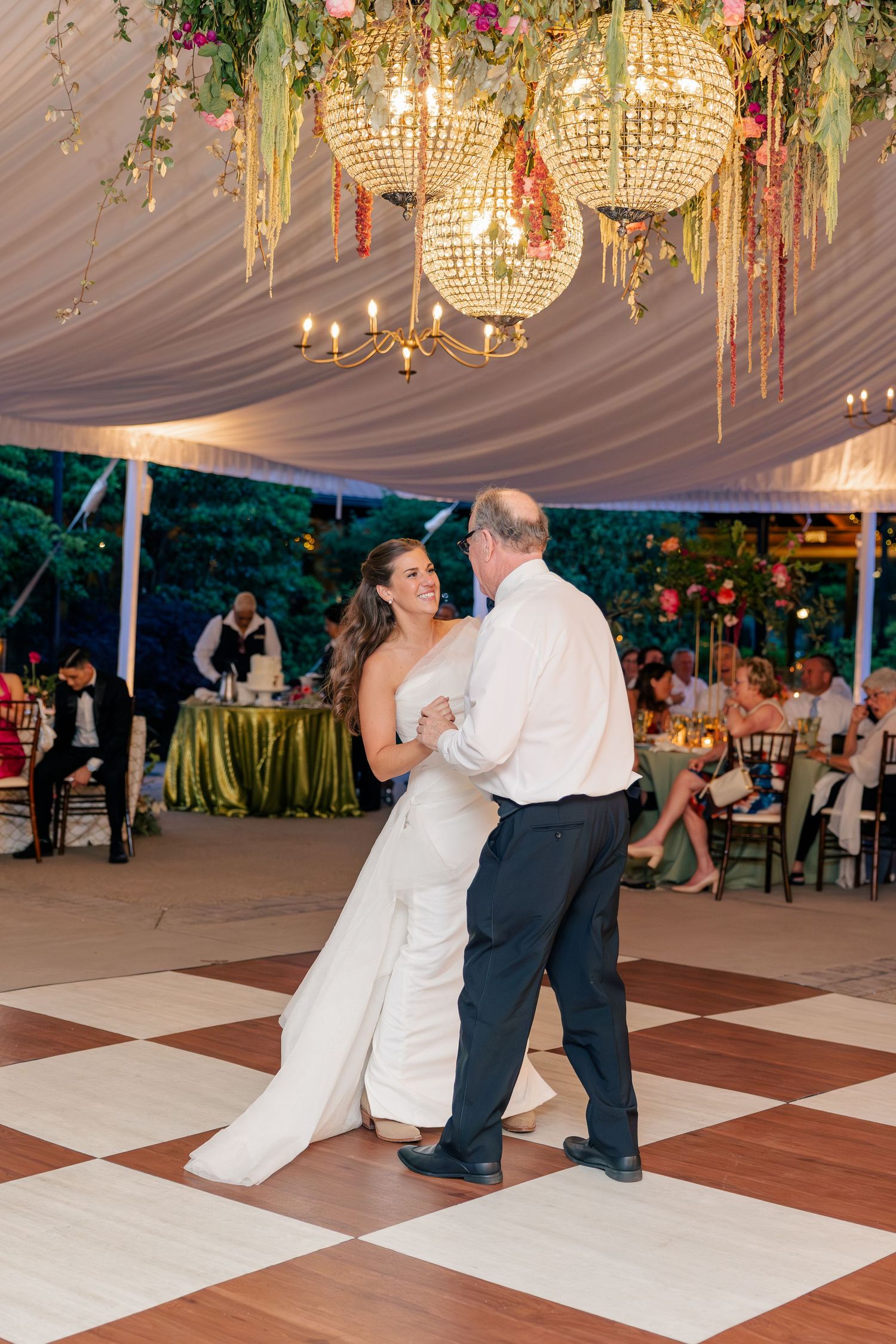 Bride dances with father under glowing chandeliers on checkered dance floor at evening reception.