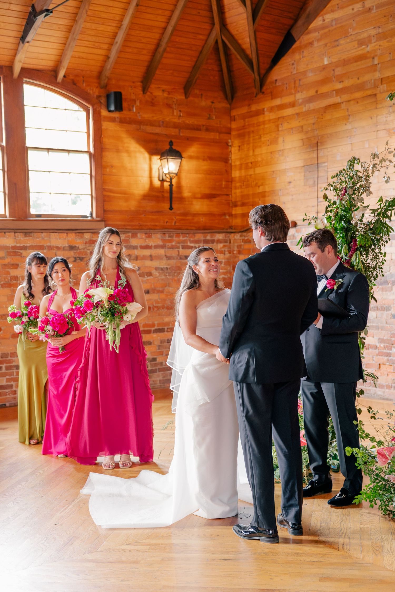 Bride and groom exchange vows at indoor wedding ceremony with bridesmaids in pink dresses and brick wall backdrop.