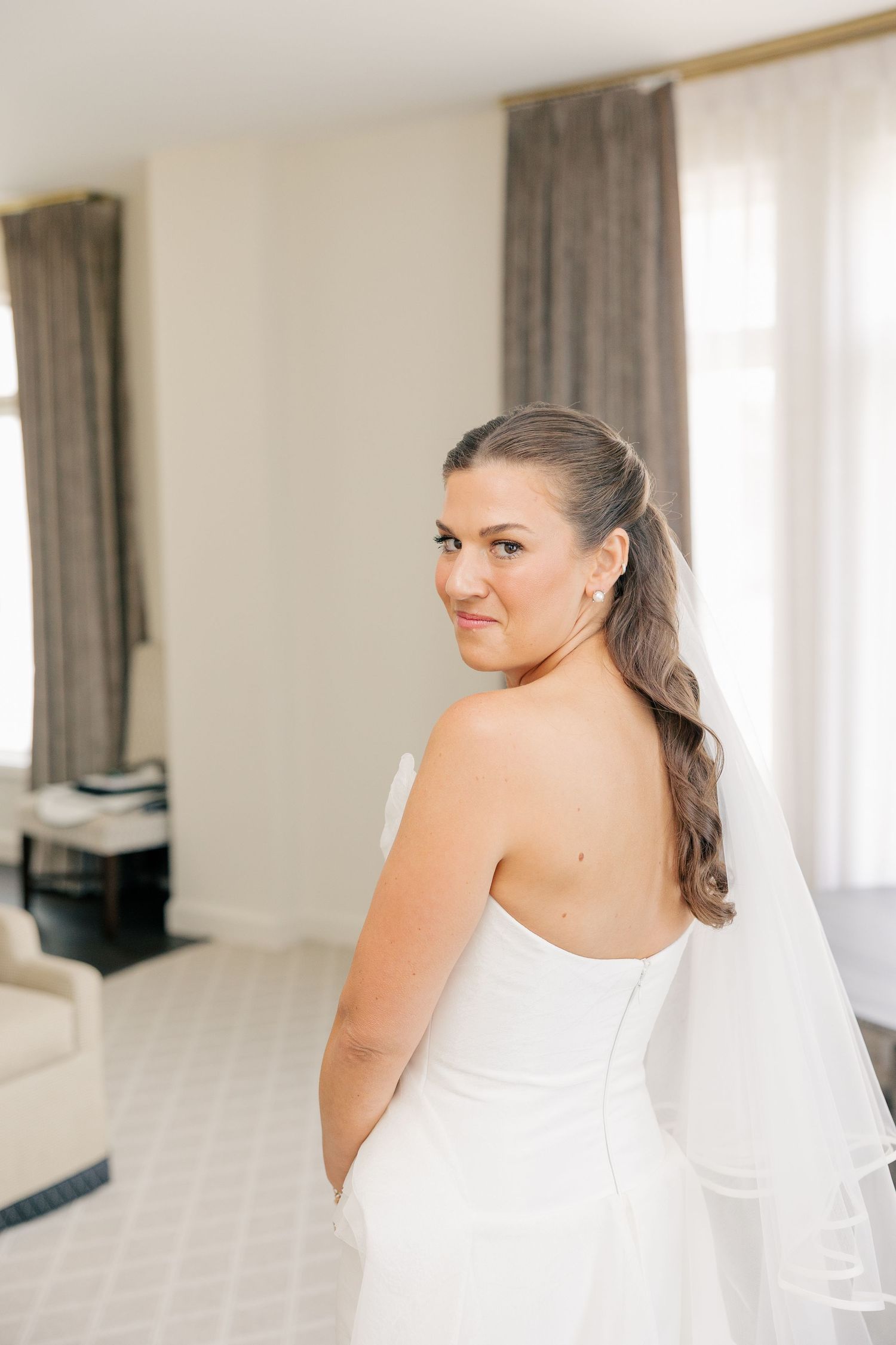 Bride in strapless backless white gown with veil glances over shoulder in bright hotel room.