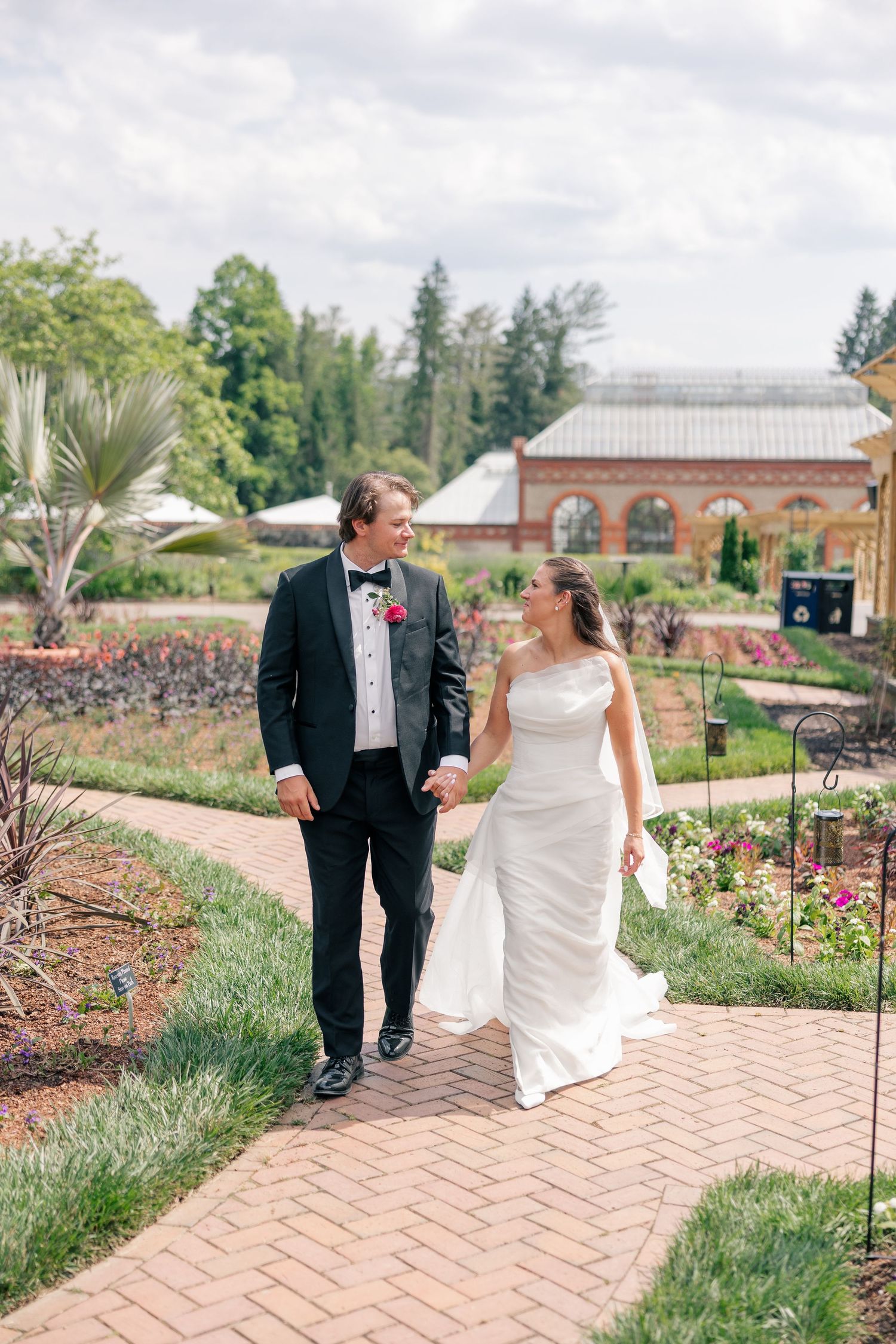 Bride and groom walk hand in hand through a colorful garden path at their outdoor wedding venue.