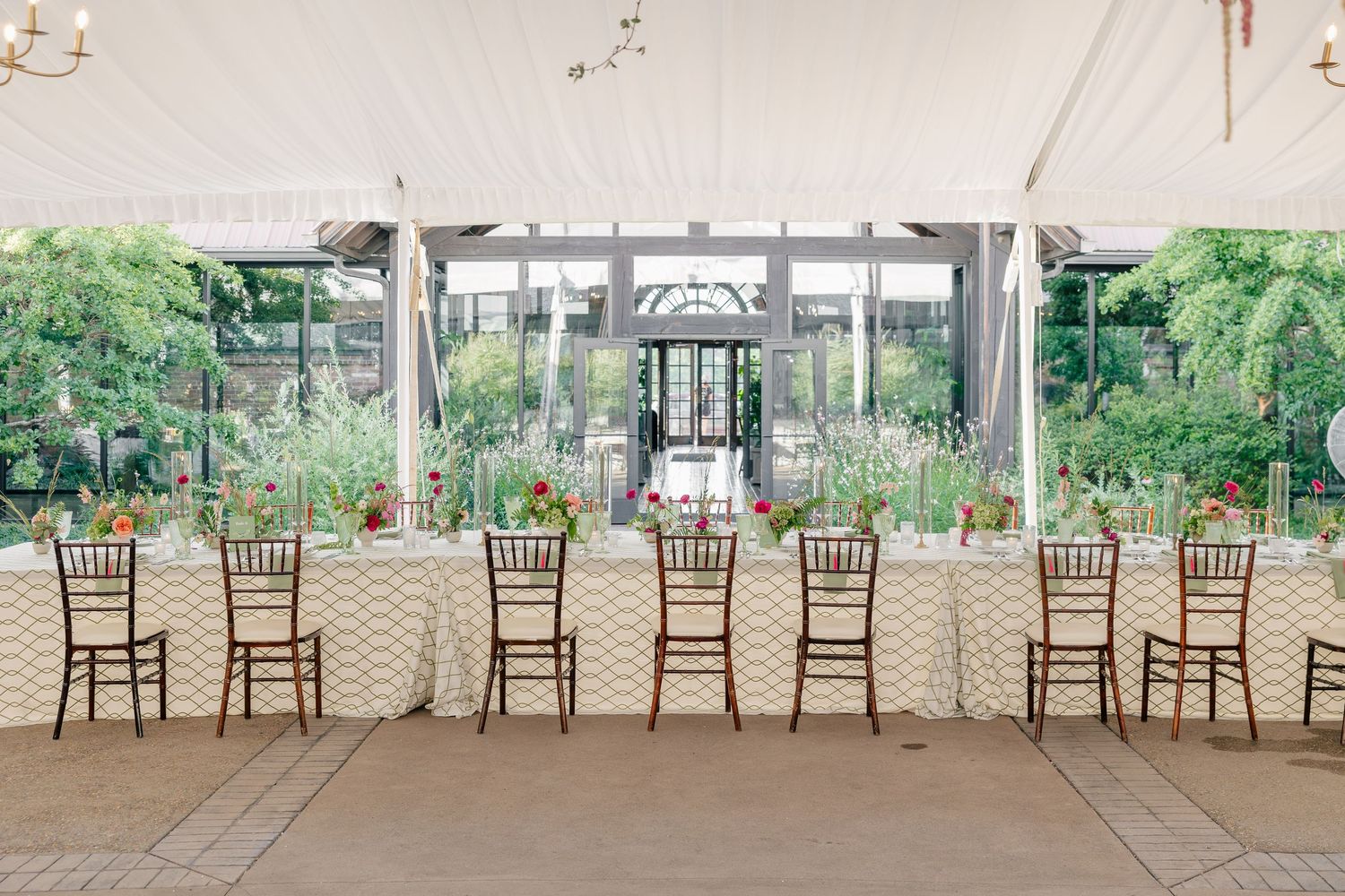 Long wedding reception table with chiavari chairs set under white tent with garden backdrop.