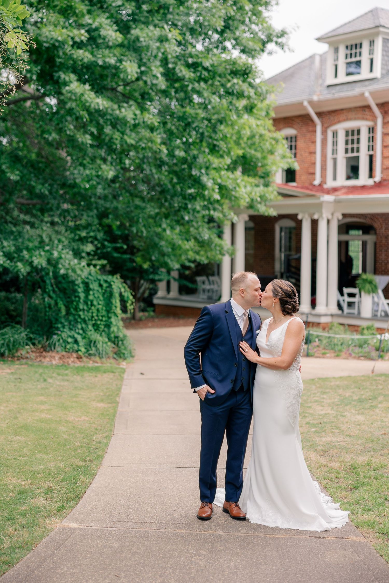 Bride and groom share a kiss on a garden path outside a historic red brick mansion on their wedding day.