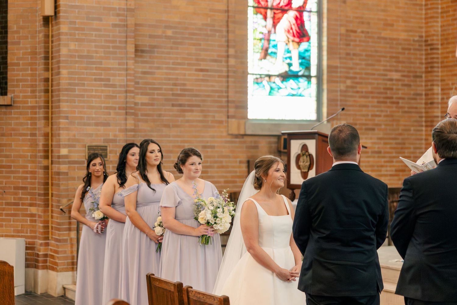 Bridesmaids in lavender dresses stand with bride at church altar ceremony.