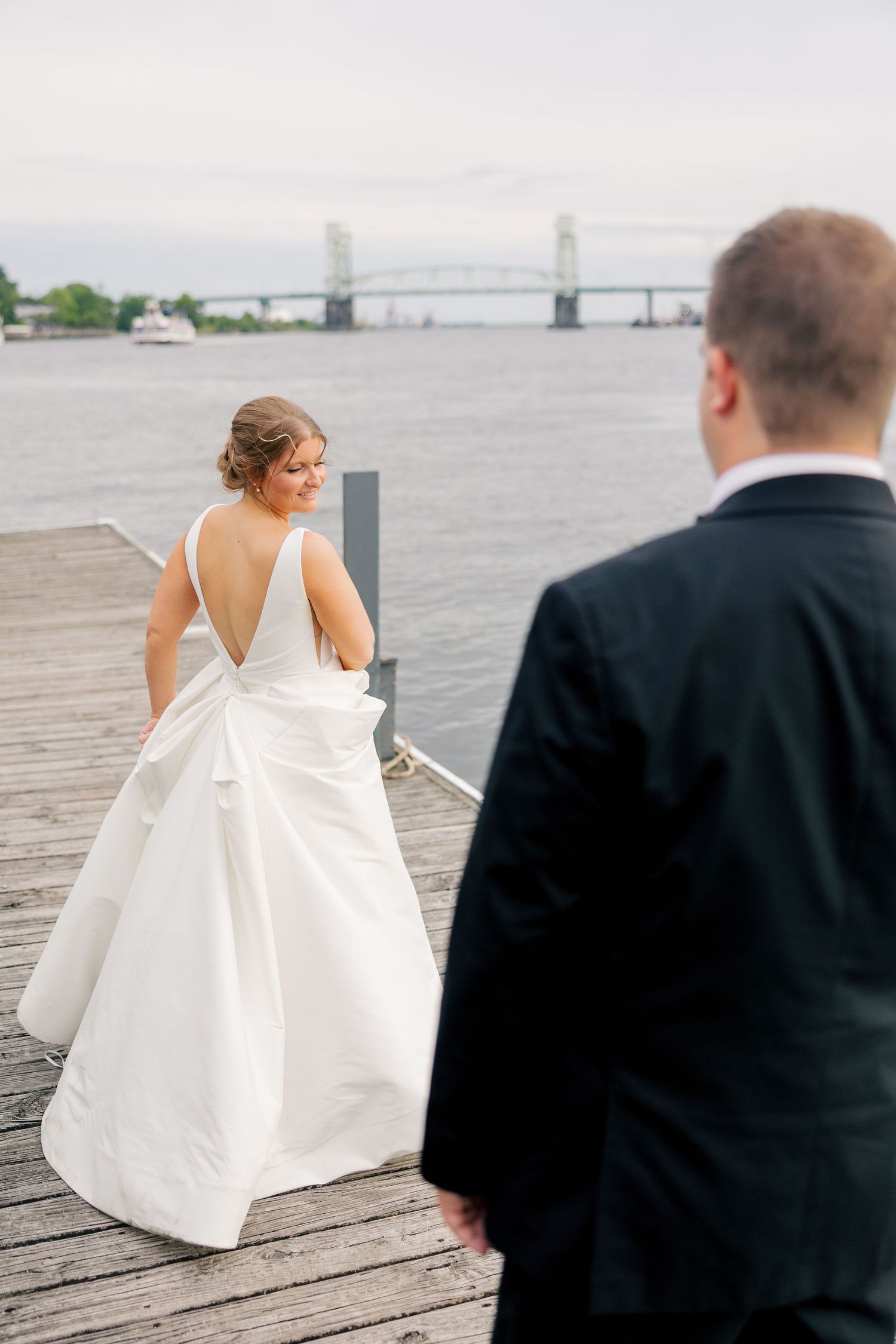 Bride in elegant backless white gown smiles at groom on scenic waterfront dock with bridge view.