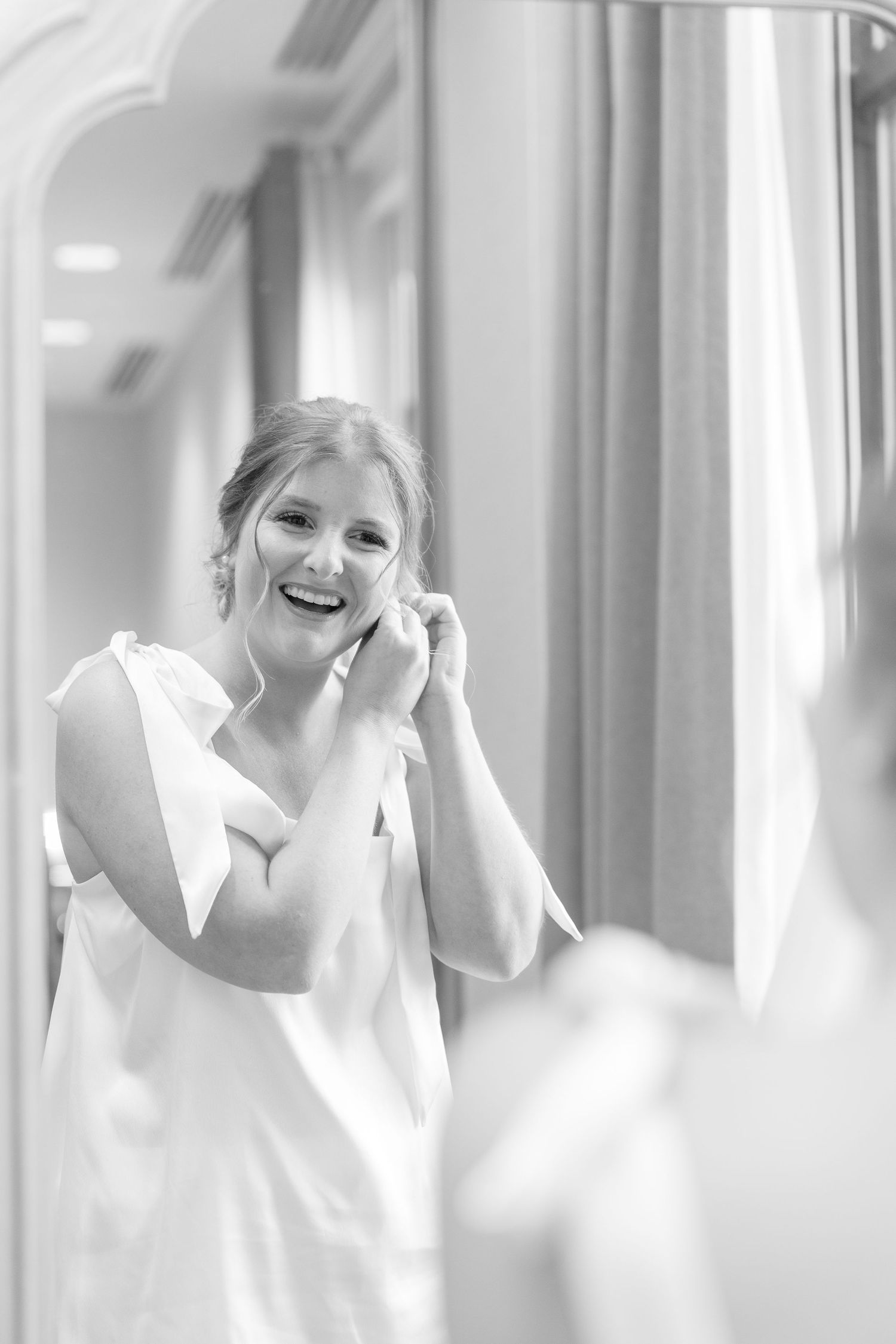 Black and white photo of smiling bride putting on earrings while getting ready in front of a mirror.
