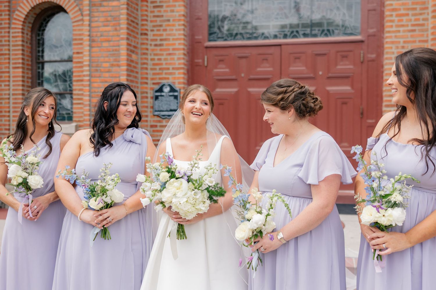 Bride laughing with four bridesmaids in lavender dresses holding white floral bouquets.