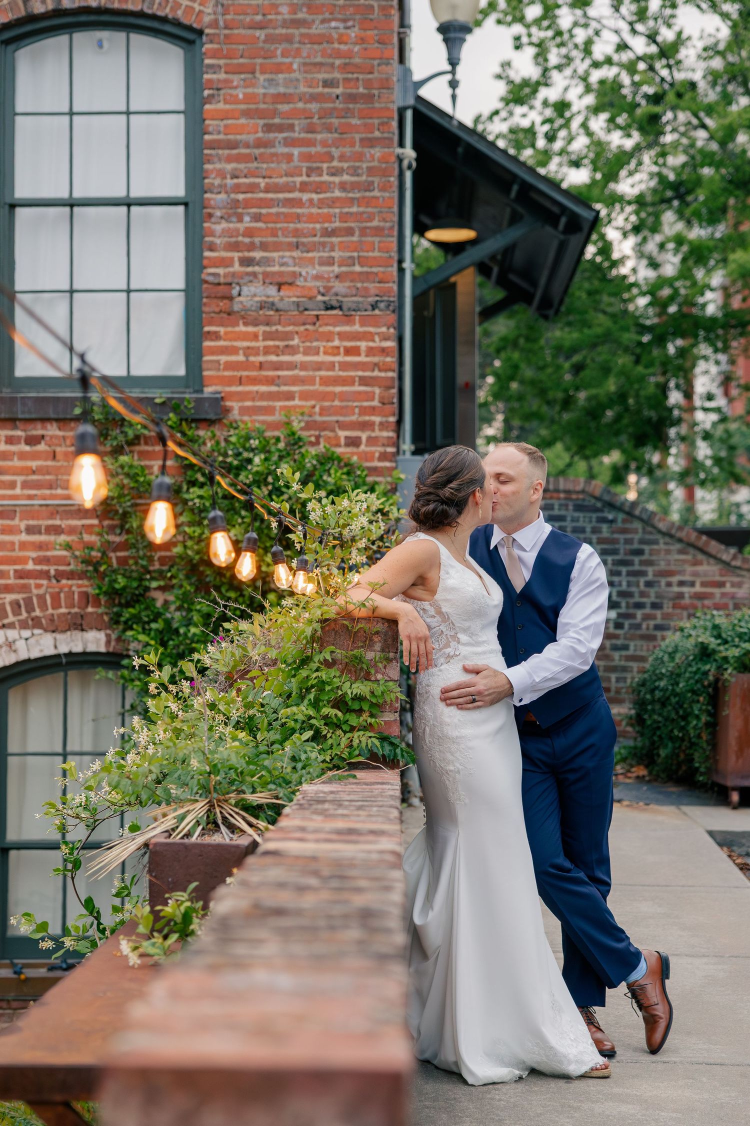 Bride and groom kissing outdoors beside brick building with string lights and lush greenery backdrop.