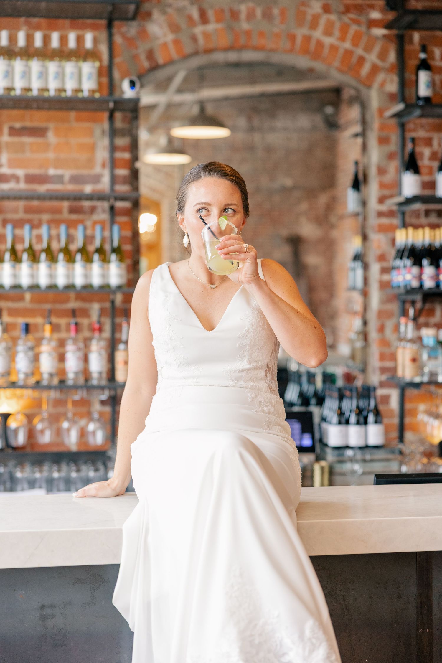 Bride in V-neck wedding gown sipping cocktail while seated at a rustic brick bar.