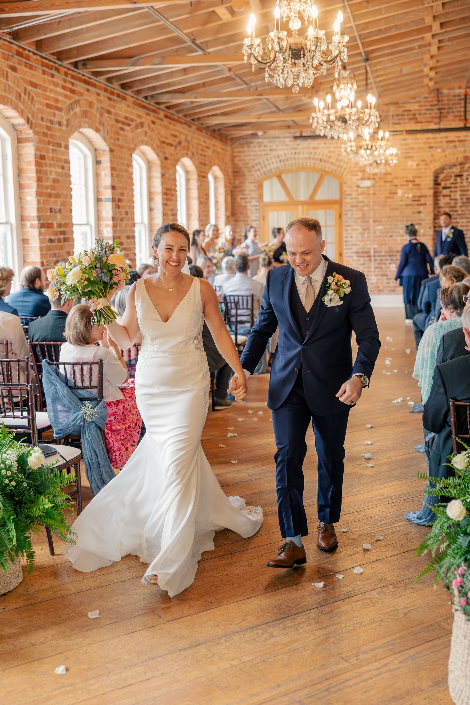 Newlyweds walk joyfully down aisle in brick-walled wedding venue after ceremony, holding hands.