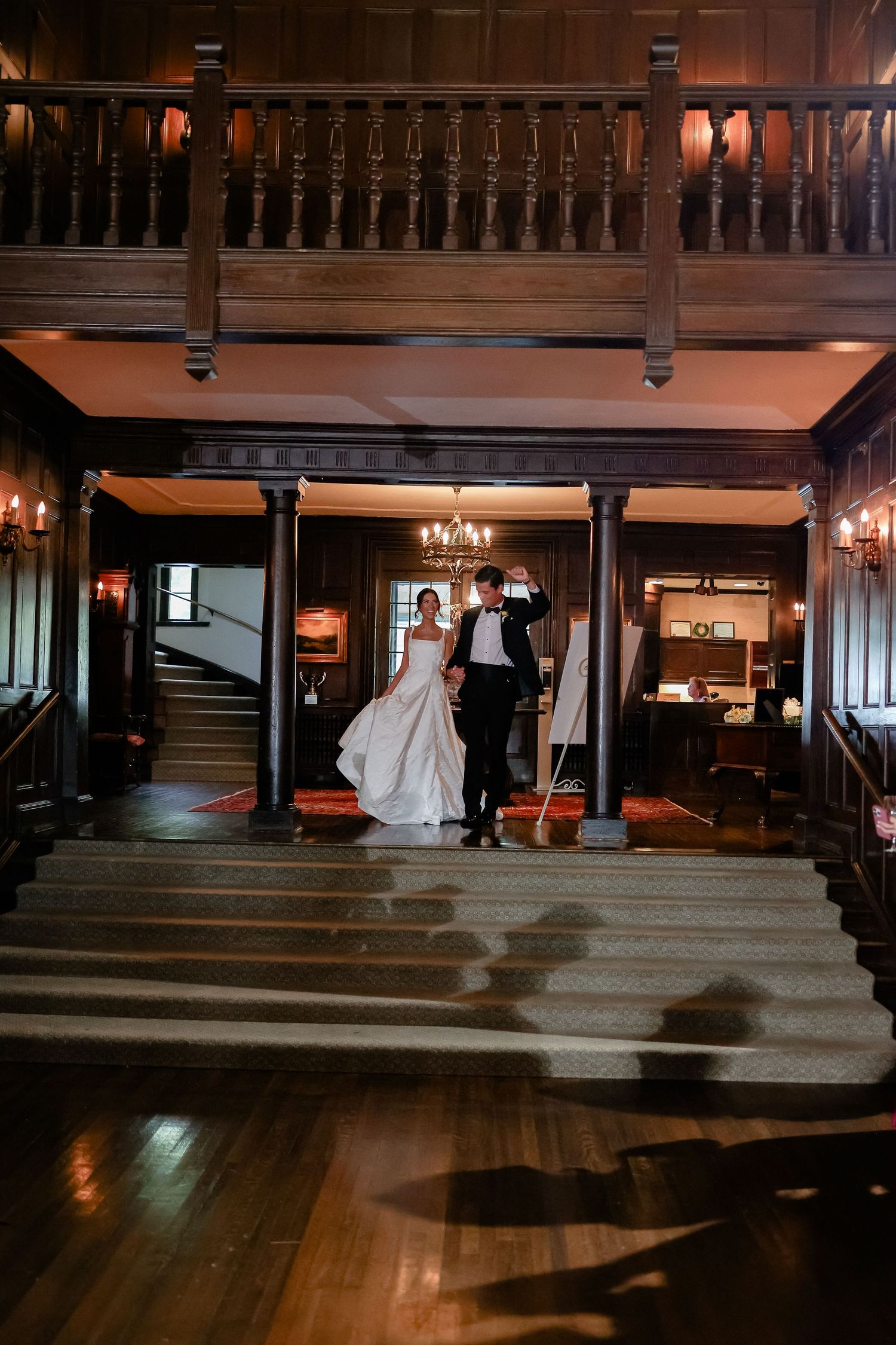 Bride and groom make grand entrance down staircase in elegant dark wood ballroom during wedding reception.