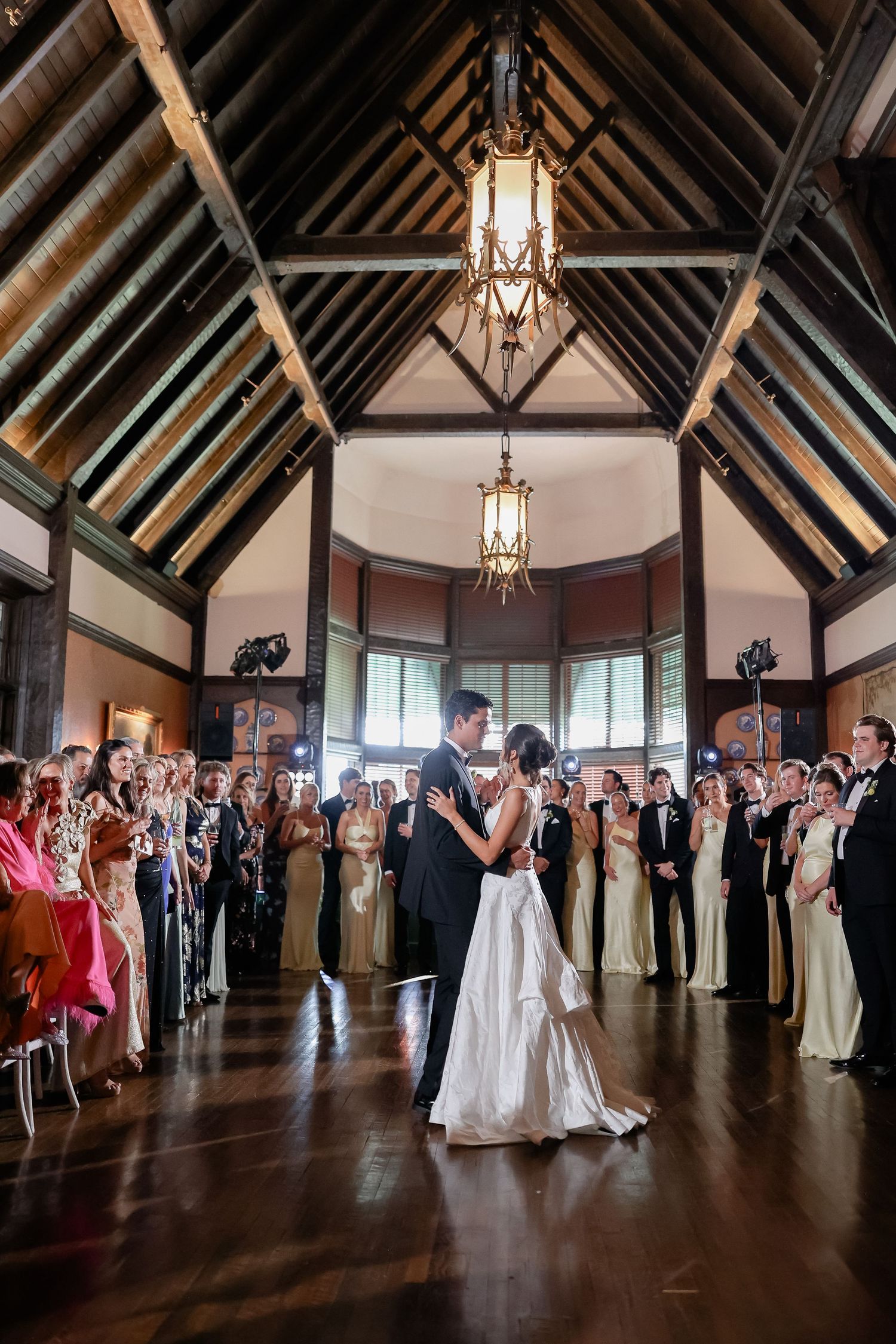 Bride and groom share their first dance in an elegant wood-beamed ballroom surrounded by wedding guests.