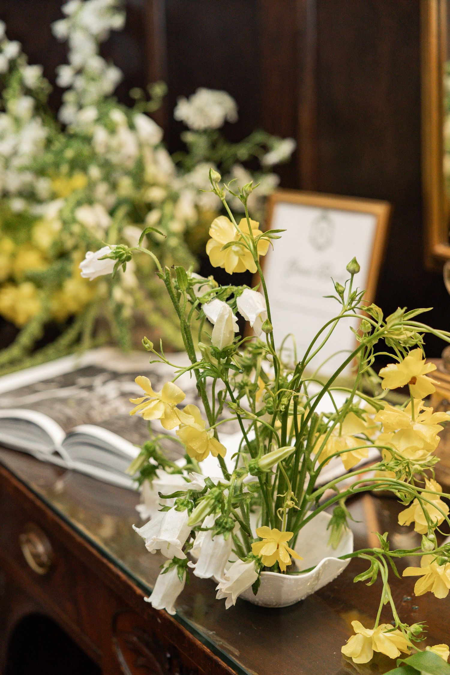 Yellow and white wildflowers arranged in a white vase on a wooden table with an open book at a wedding reception.