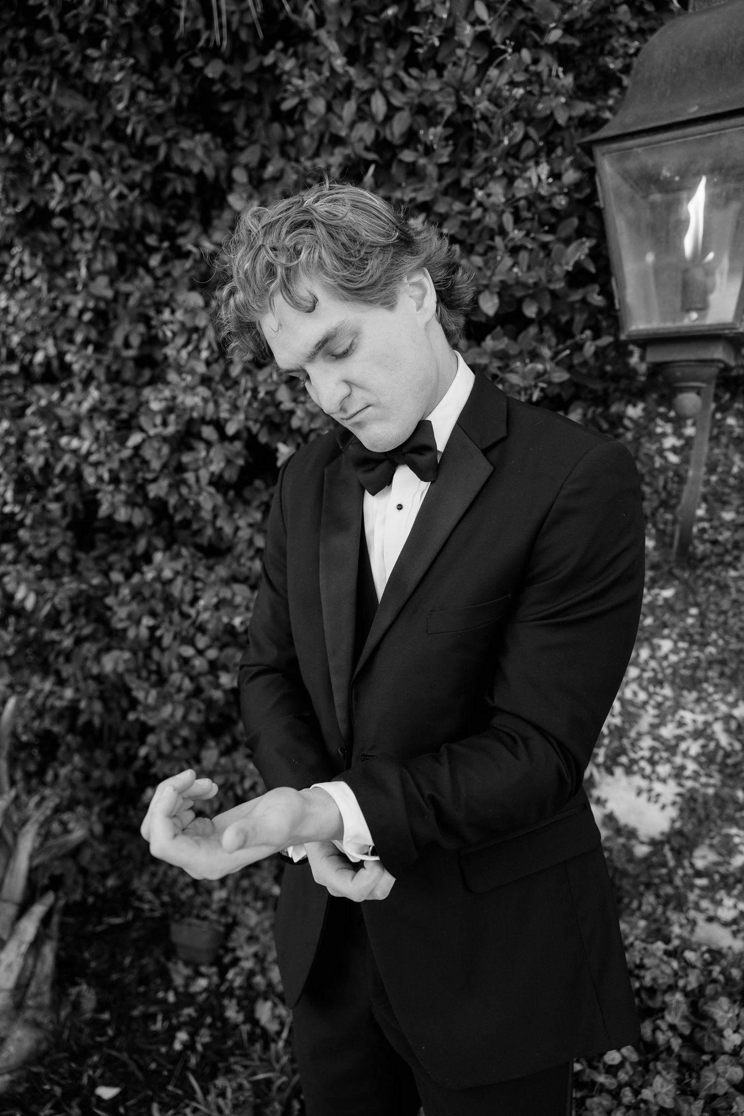 Groom in black tuxedo adjusts his cufflink beside a garden lantern in black and white portrait.