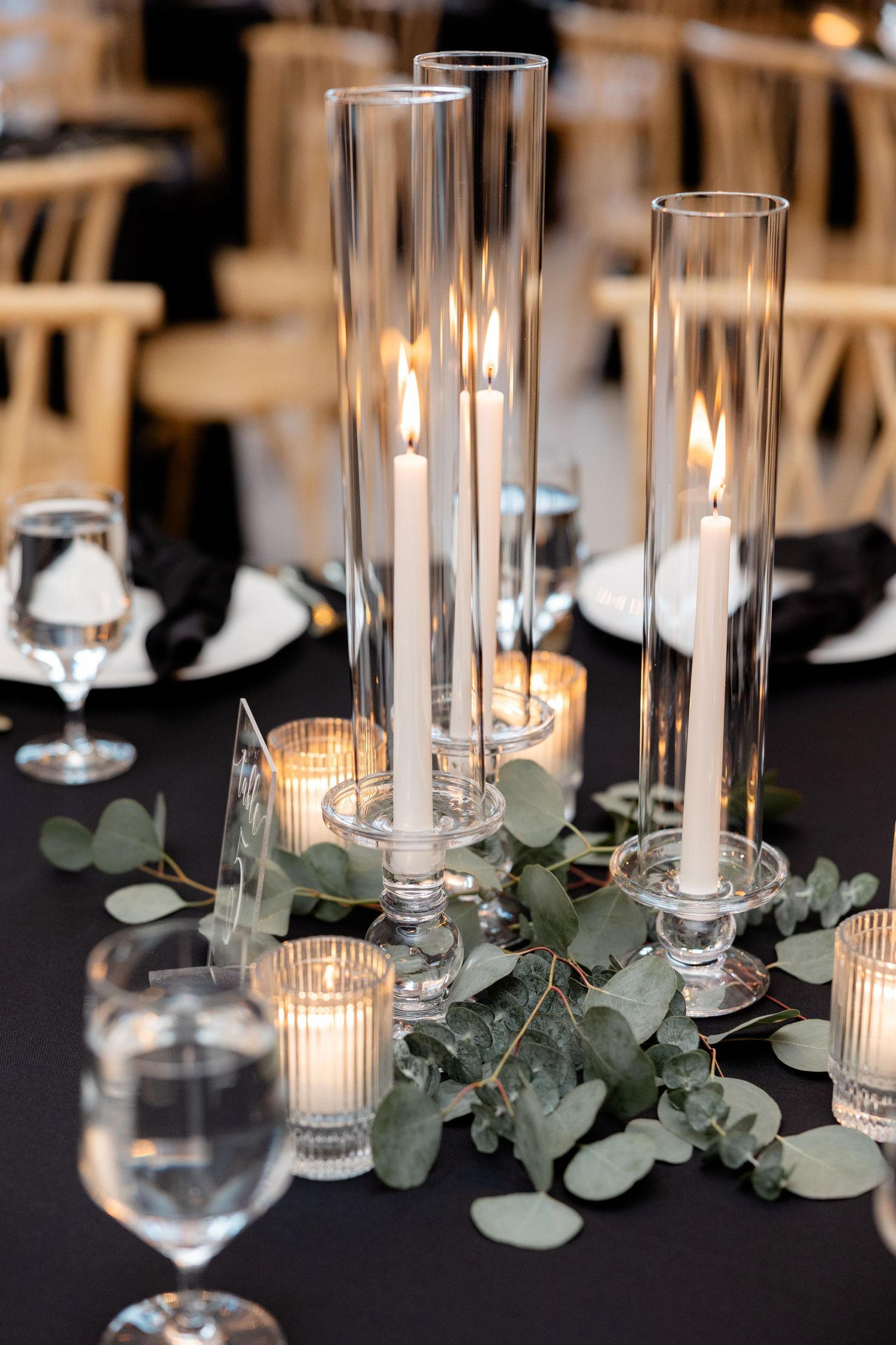 Elegant wedding table centerpiece with tall glass candle holders, eucalyptus leaves, and votive candles.