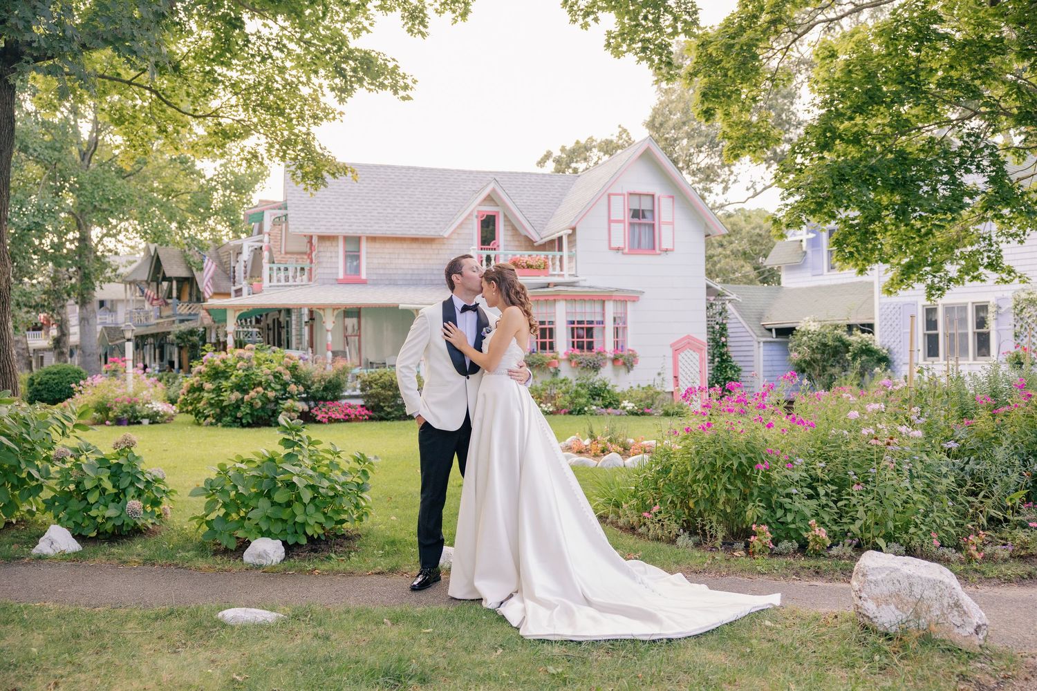 Bride and groom share a romantic kiss in front of a charming pink Victorian house surrounded by lush gardens.