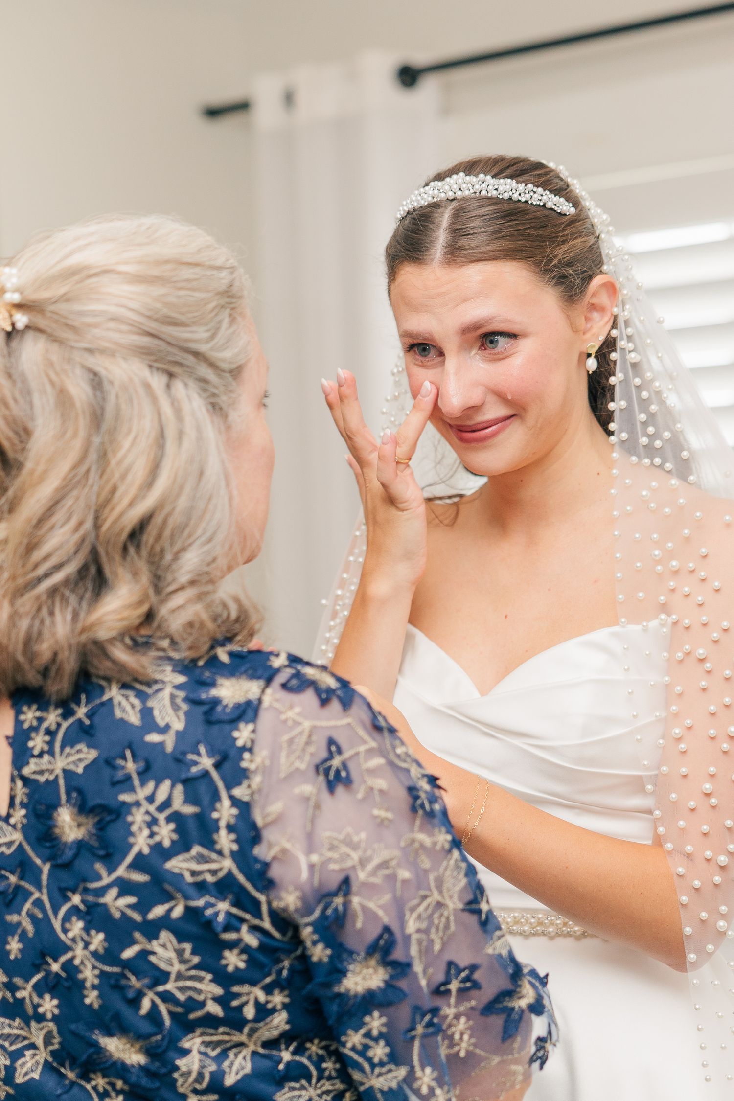 A bride wipes tears as an older woman in a blue floral dress tenderly touches her face before the wedding ceremony.