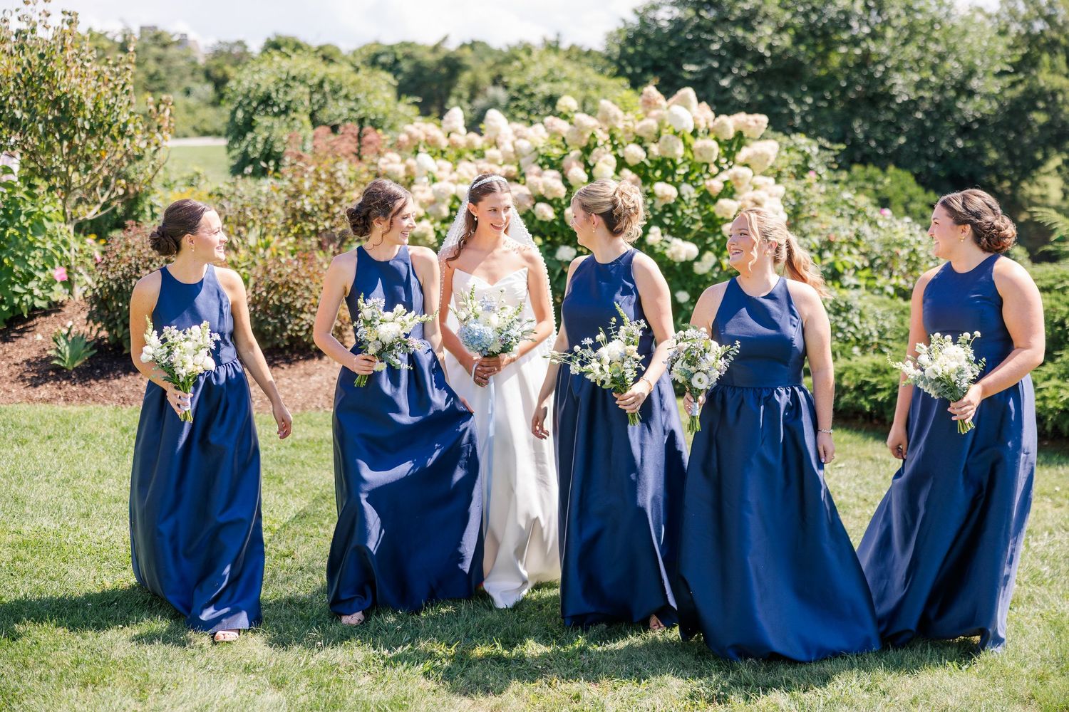 Bride with bridesmaids in navy blue gowns holding white bouquets in a garden setting.