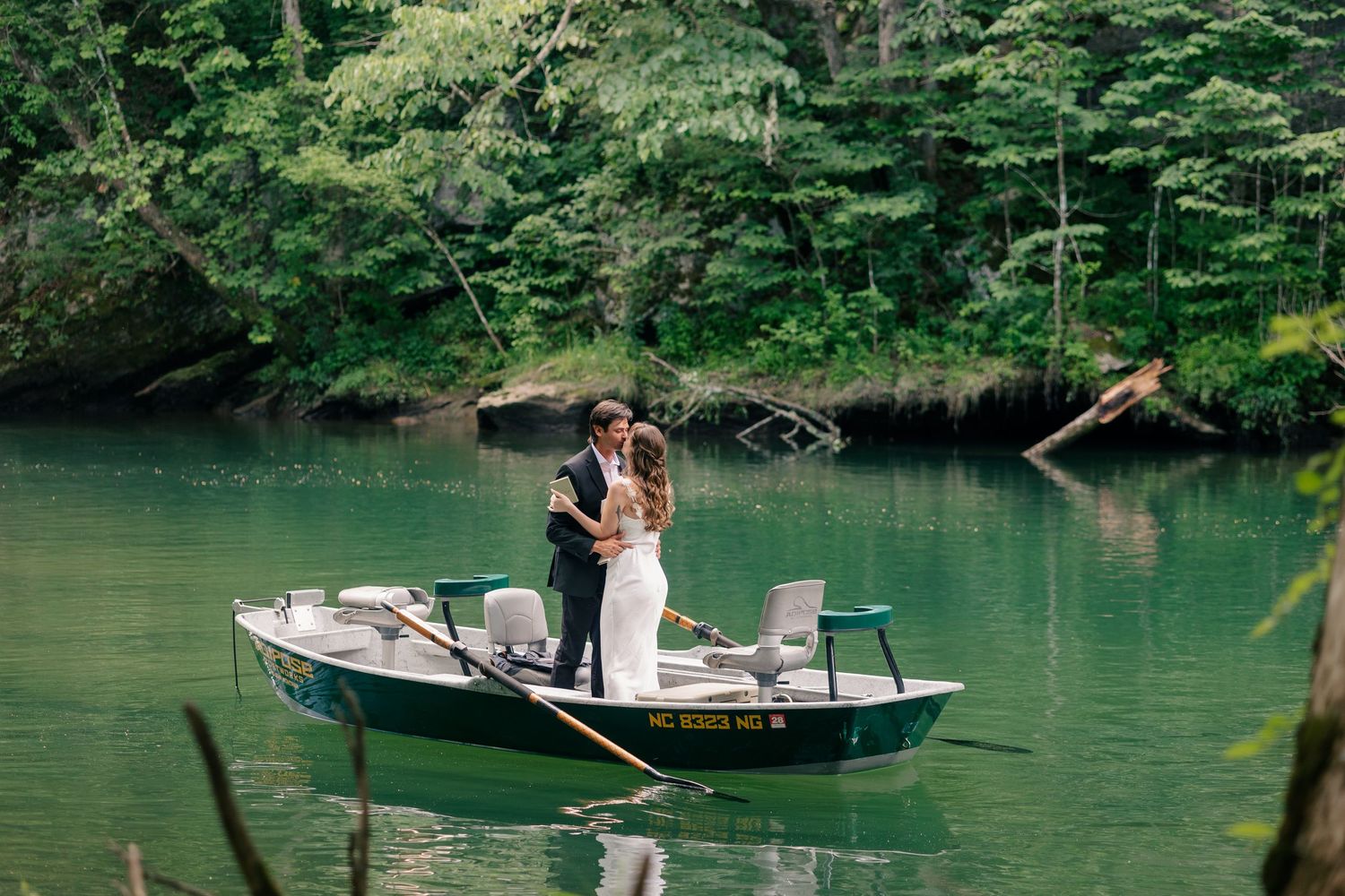 Bride and groom share a romantic kiss standing in a green rowboat on a serene lake surrounded by lush forest.