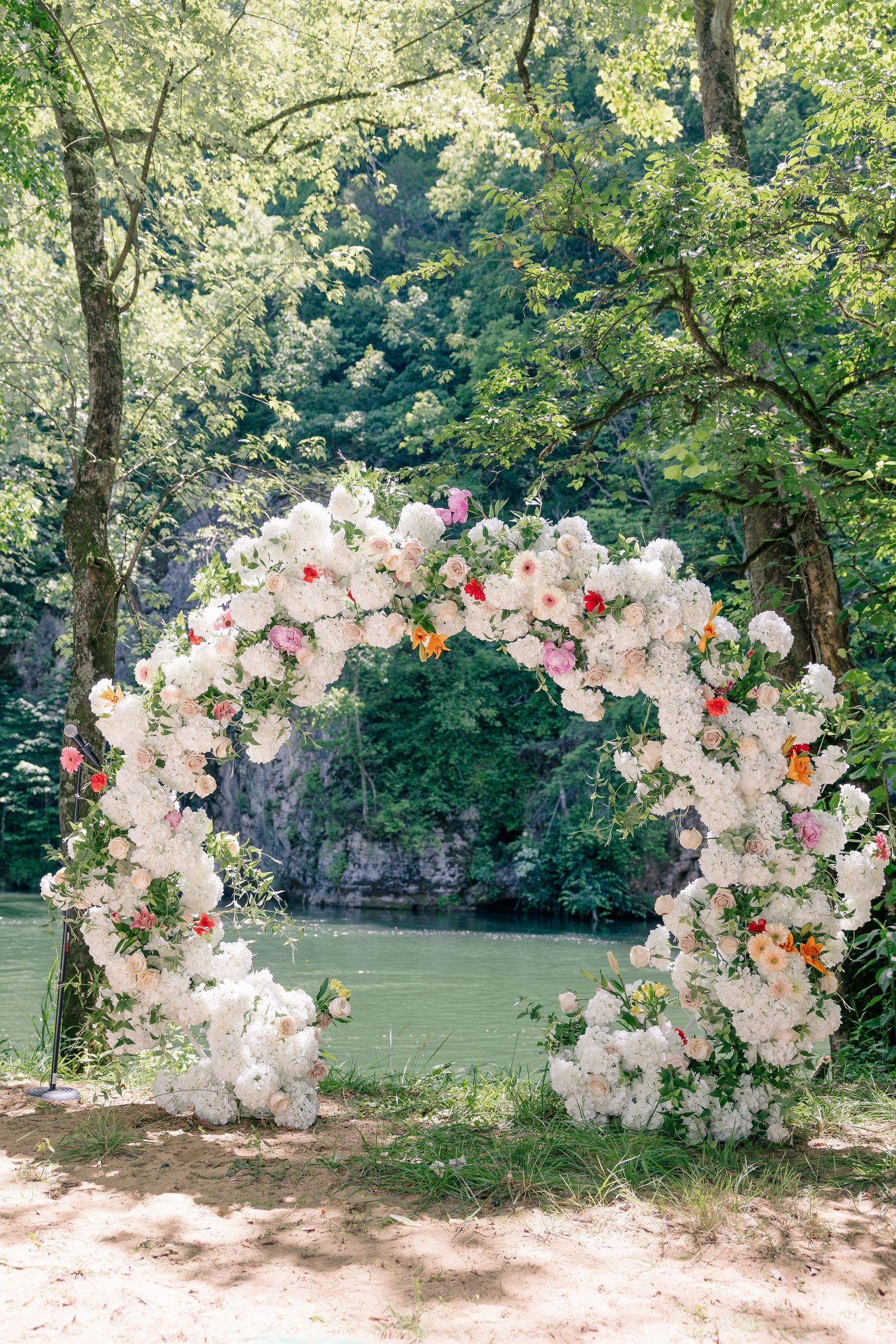 Circular floral wedding arch adorned with white and colorful blooms beside a serene green river.