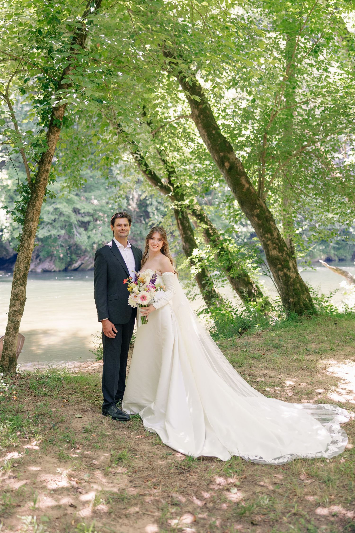 Wedding couple posing under green trees by a river, bride in a long white gown with train holding a floral bouquet.