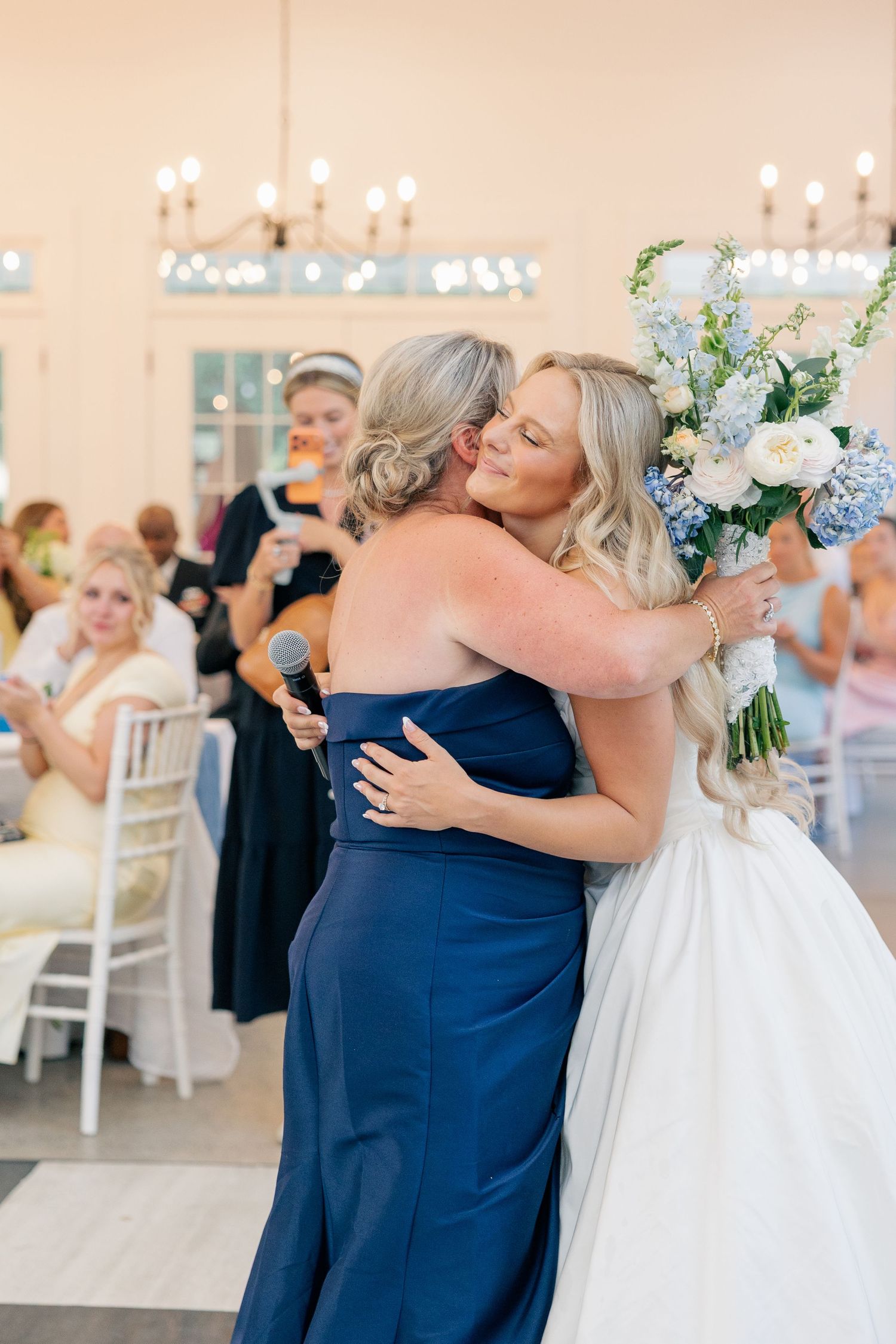 Bride in white gown hugging woman in navy dress holding blue and white floral bouquet.