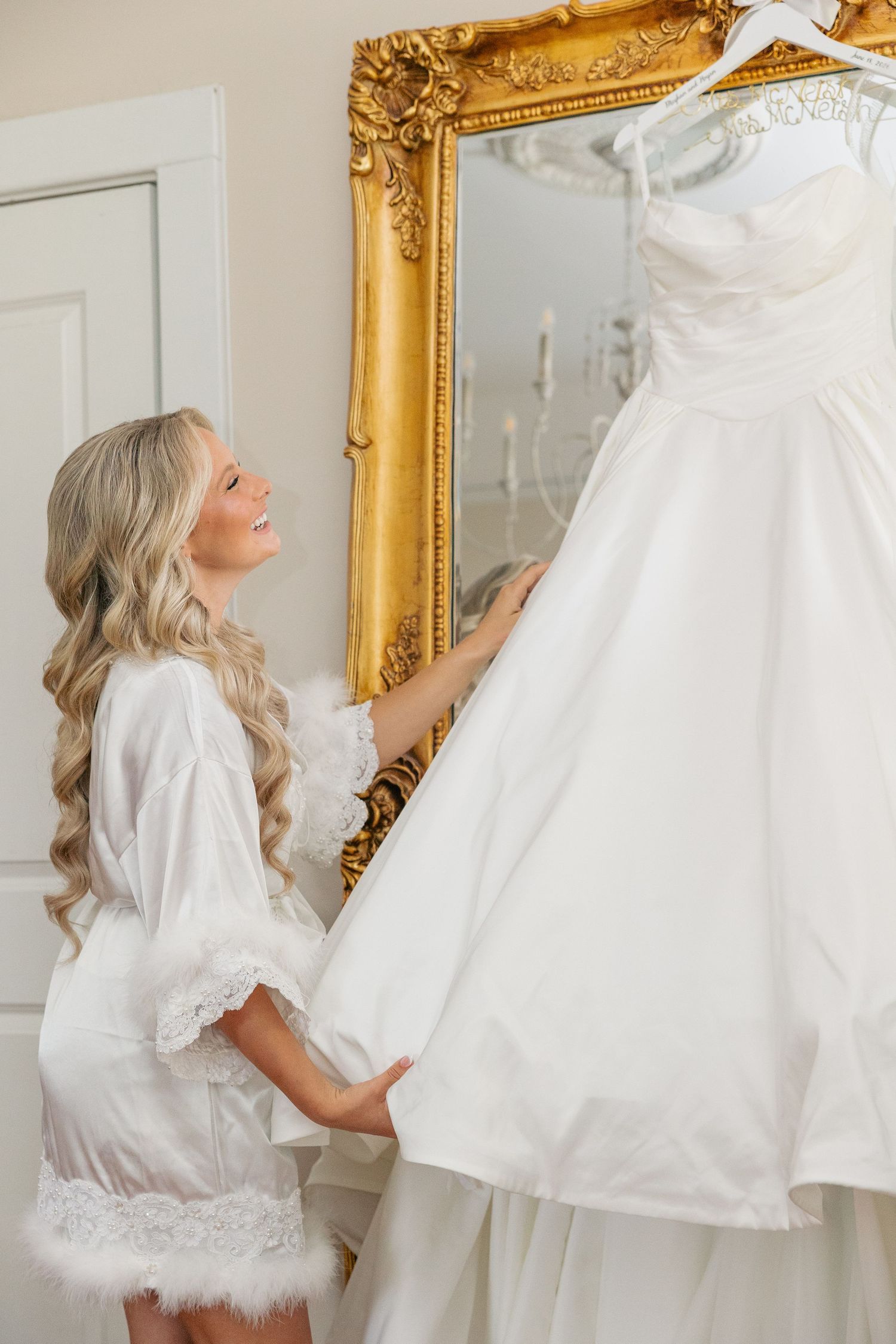 Smiling bride in white robe looking up at her wedding dress hanging by a gold mirror.