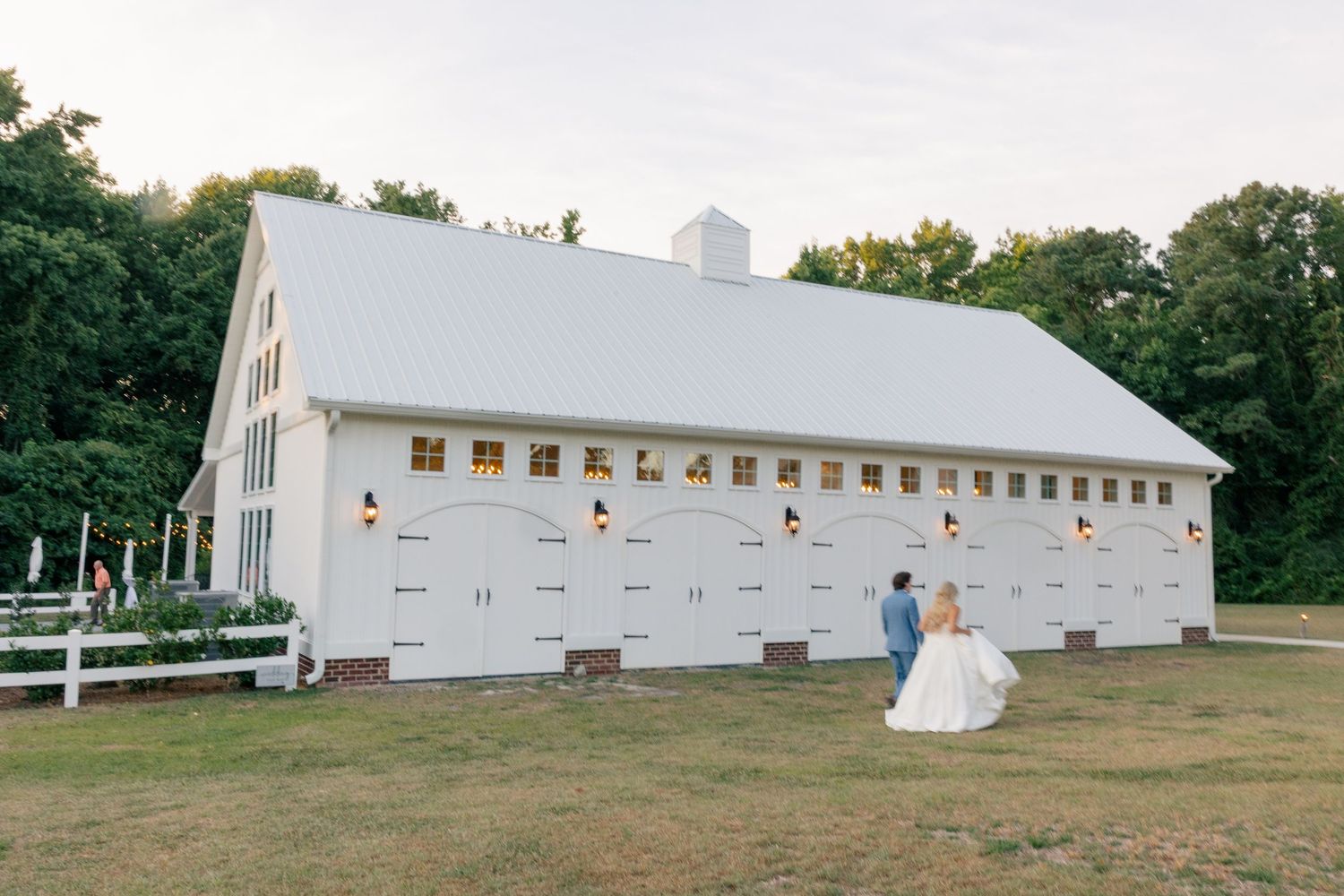 Bride and groom walk hand in hand toward large white barn wedding venue at dusk.