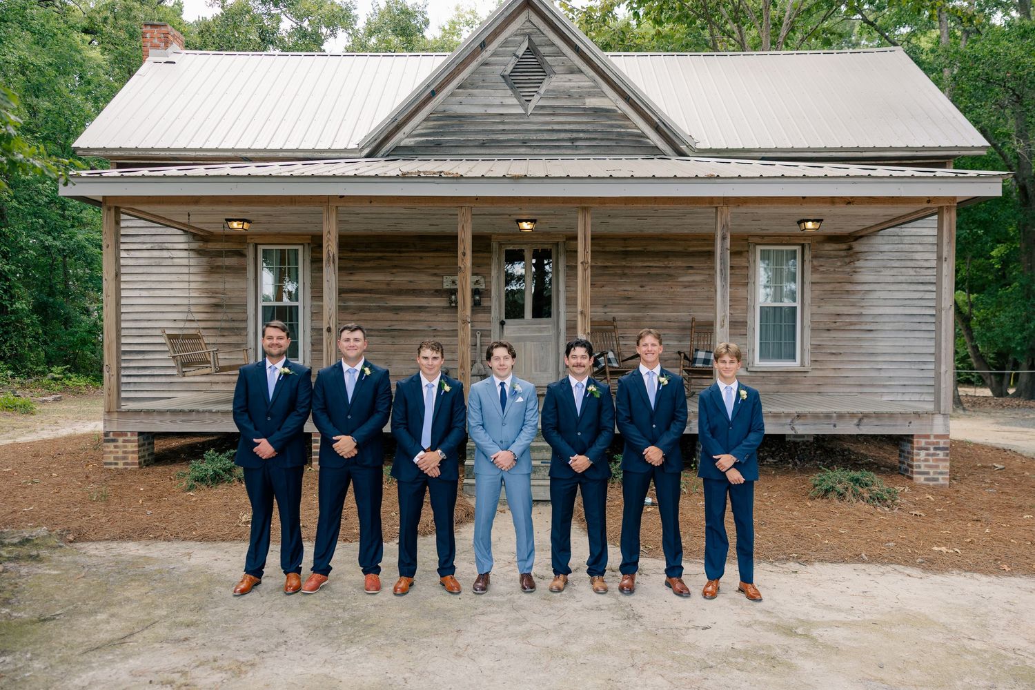 Groom in light blue suit stands with six groomsmen in navy suits in front of a rustic wooden farmhouse.