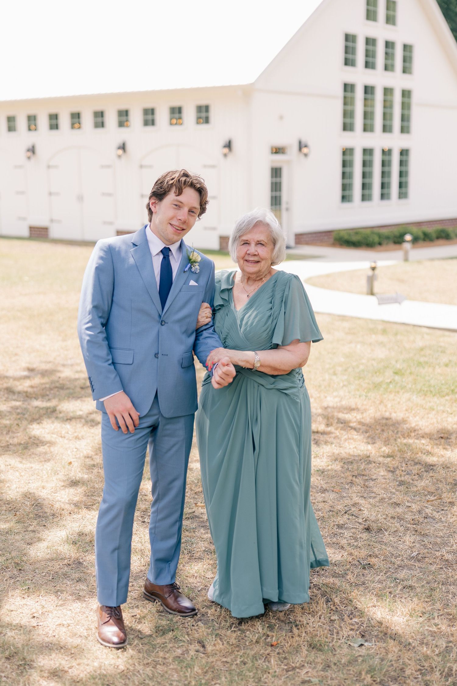 Groom in light blue suit escorts elderly woman in sage green dress at outdoor wedding venue.