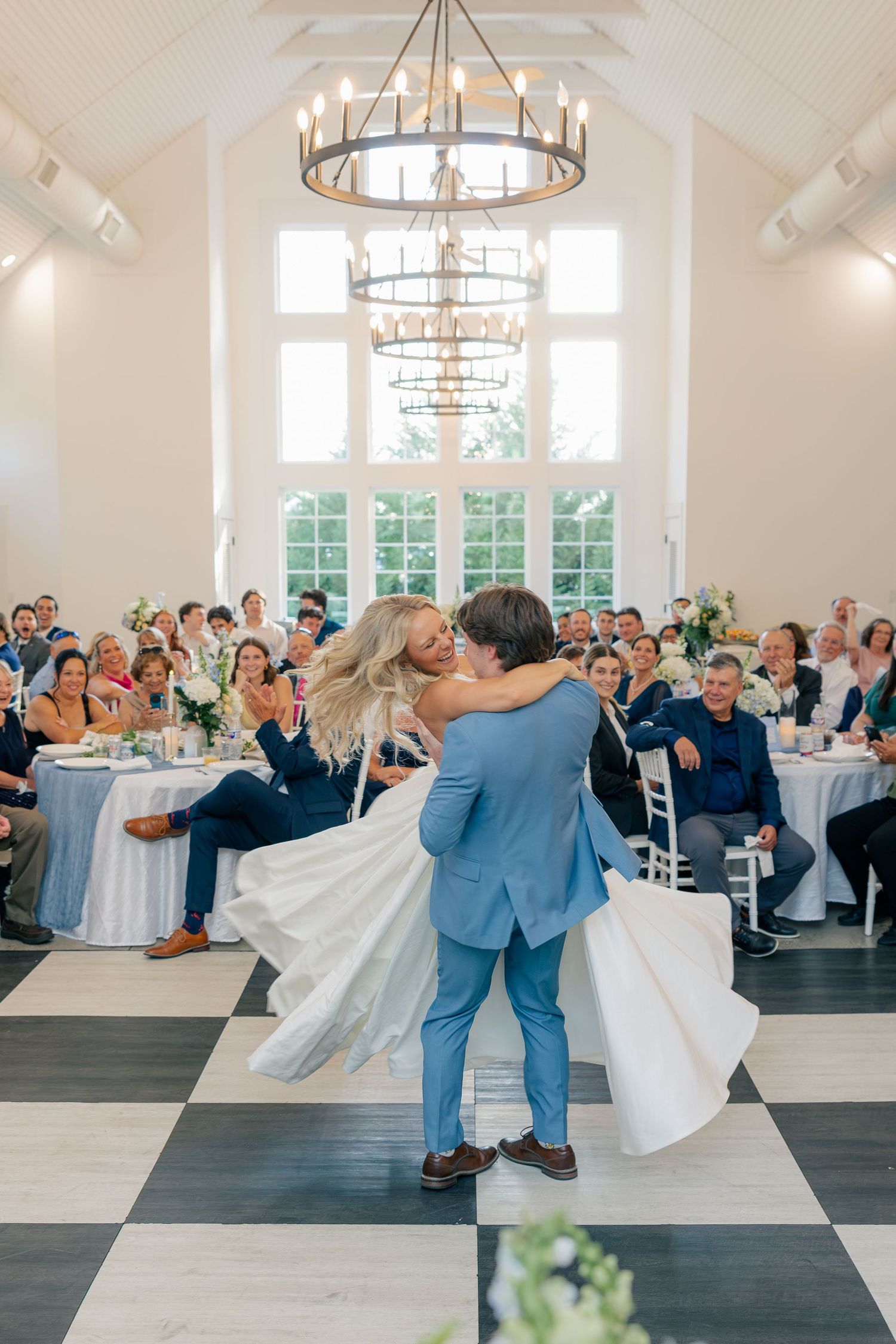 Groom lifts bride during romantic first dance on checkered floor in elegant bright reception hall.