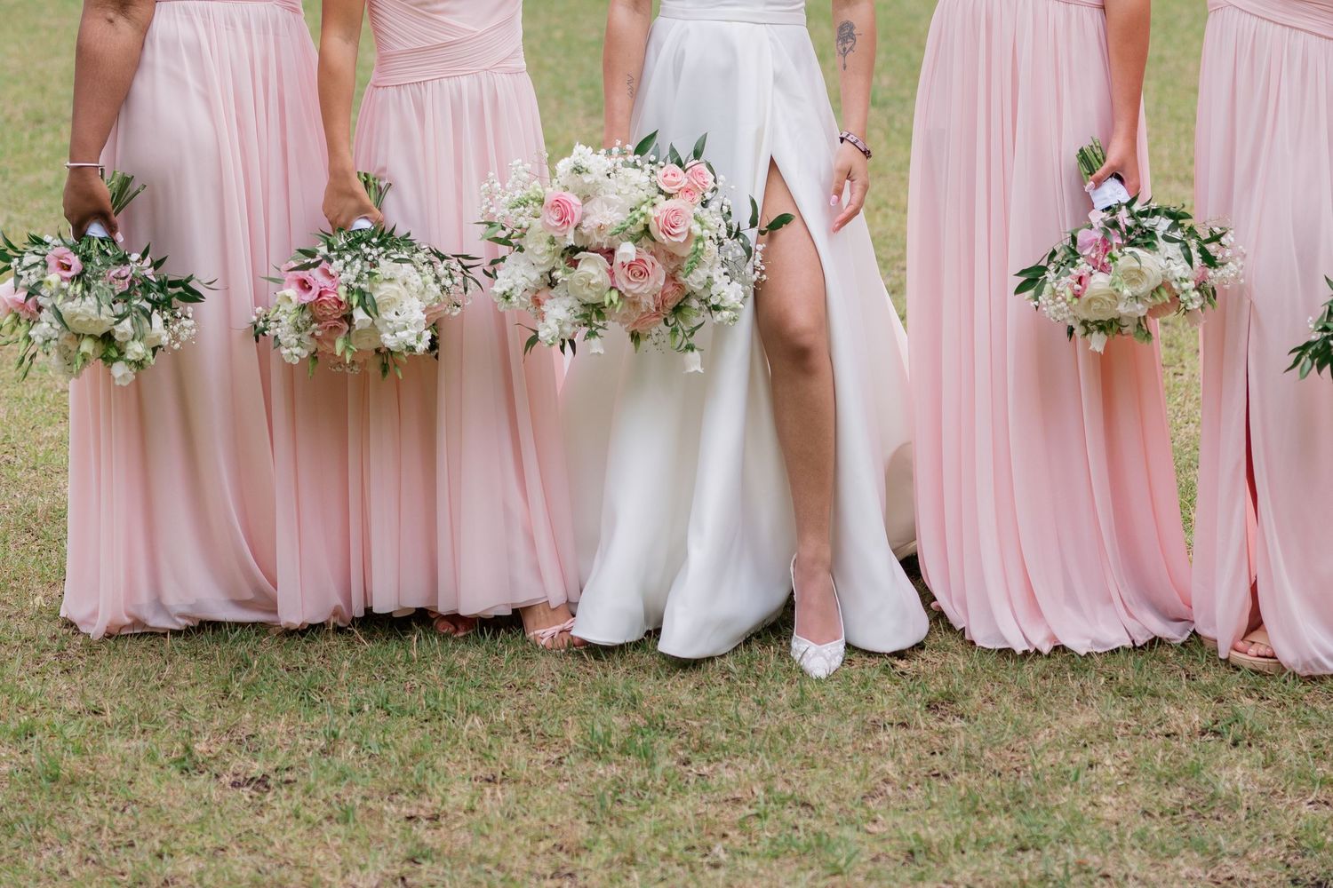 Bridesmaids in blush pink dresses lined up outdoors holding pastel floral bouquets.