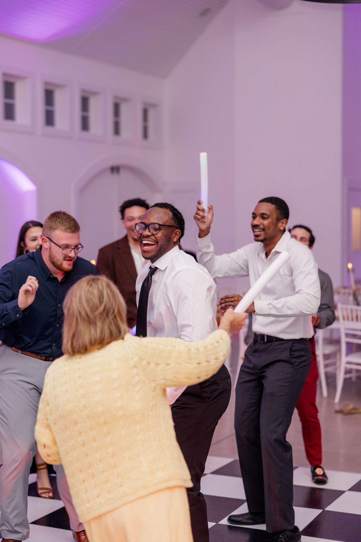 Wedding guests dancing and celebrating energetically under purple lighting.