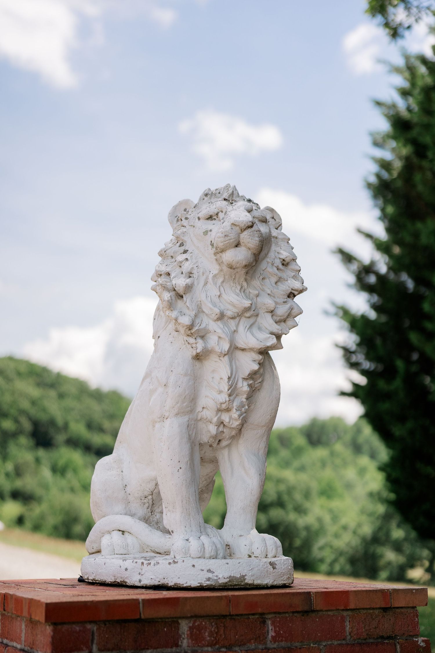 A decorative stone lion statue sits atop a brick pedestal against a backdrop of green trees and blue sky.