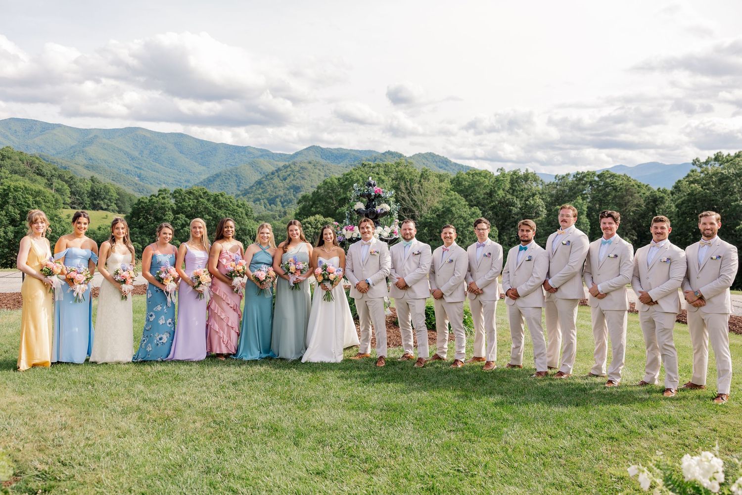 Wedding party members in pastel dresses and light suits pose in a line against mountain scenery.