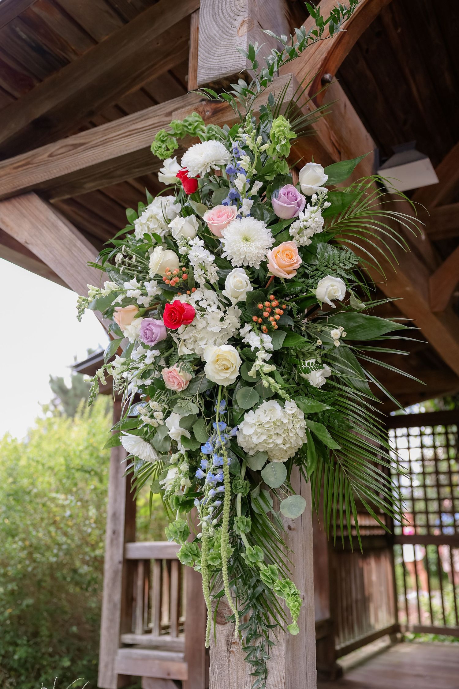 Elegant floral arrangements with white and pink roses decorate a wooden pergola for an outdoor wedding ceremony.