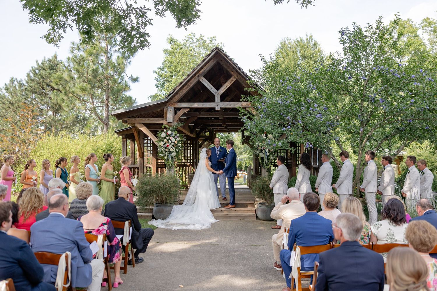 An outdoor wedding ceremony under a rustic wooden pavilion surrounded by trees and guests.