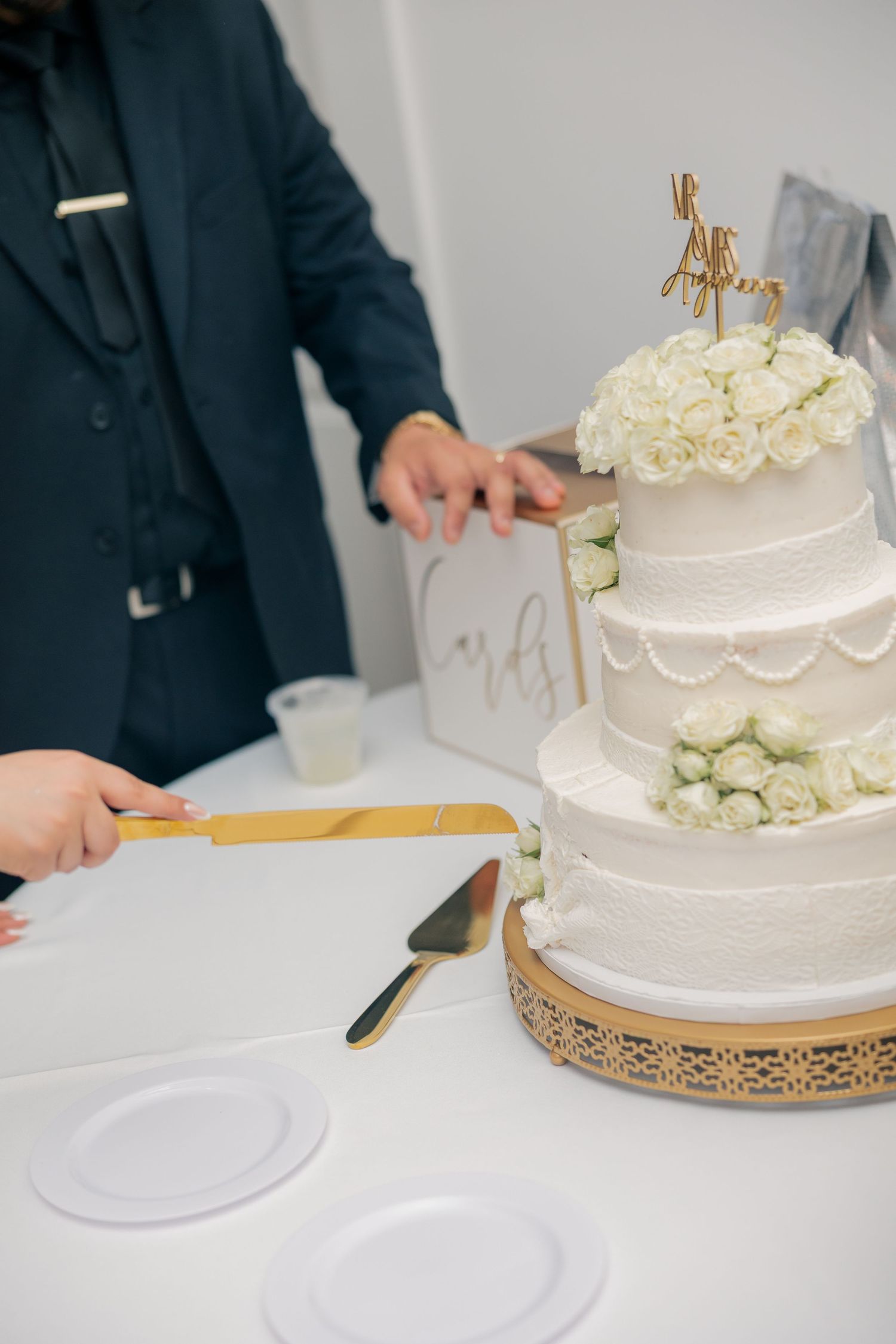 Elegant white tiered wedding cake with rose decorations being cut on white tablecloth.