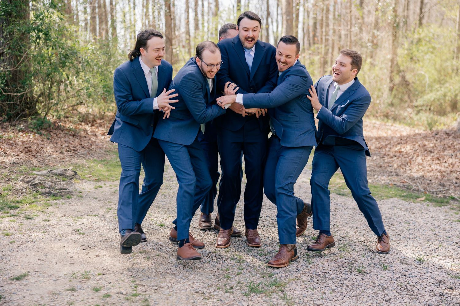 Wedding party in navy suits posing playfully together in a wooded outdoor setting.