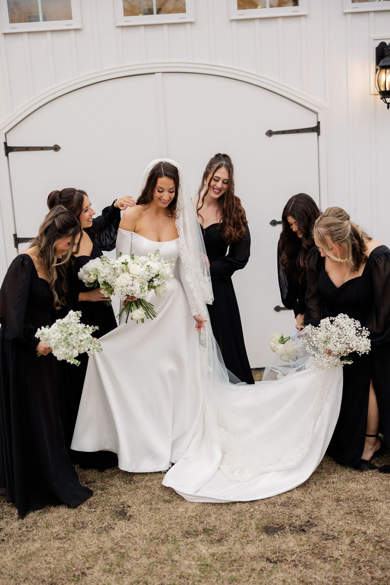 Bridesmaids in black dresses surround and support the bride in a white wedding gown against a light colored exterior wall.