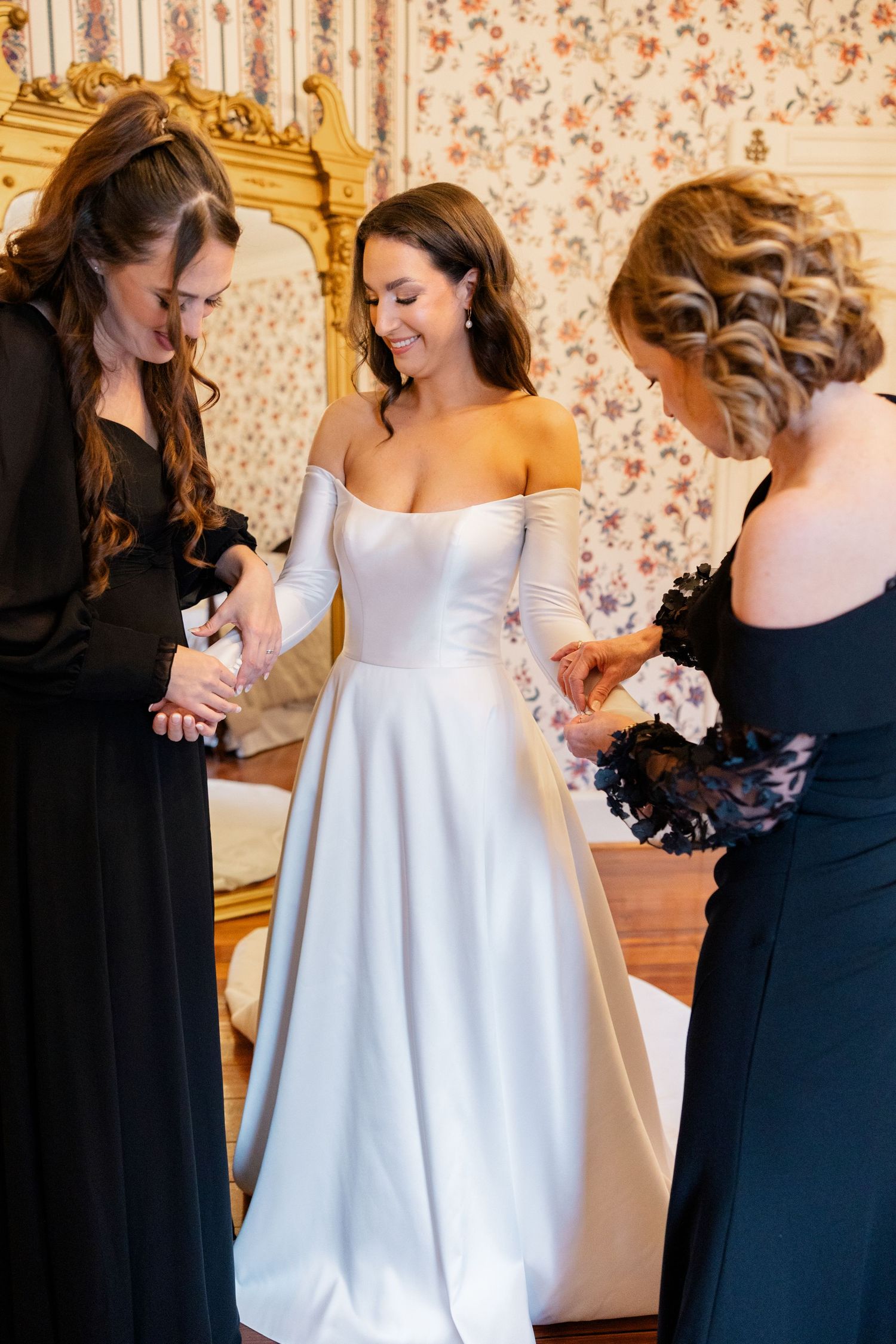 Three women sharing an emotional moment together in a room with floral wallpaper during wedding preparations.