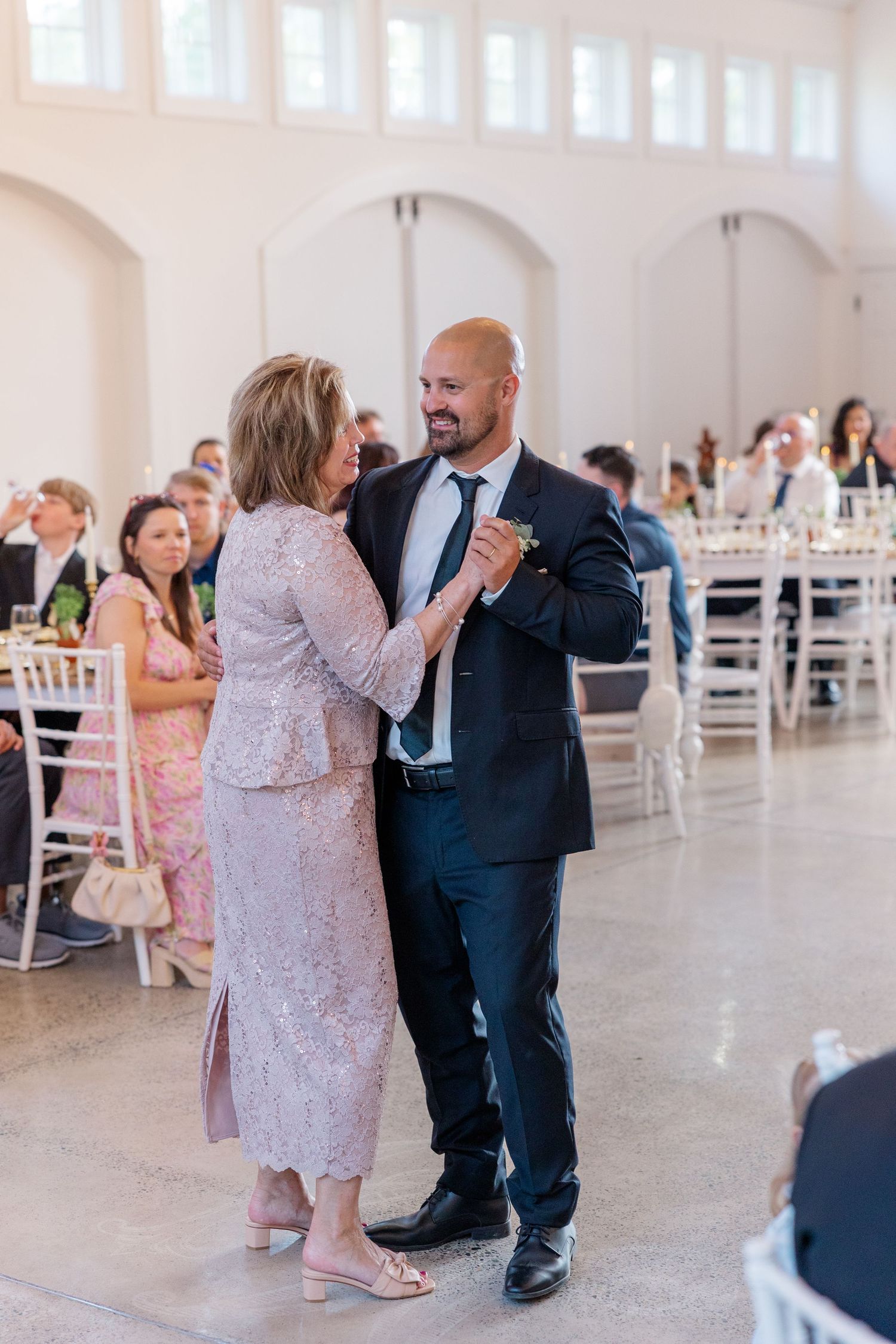 Couples dancing at an elegant wedding reception in a bright white venue with arched windows.