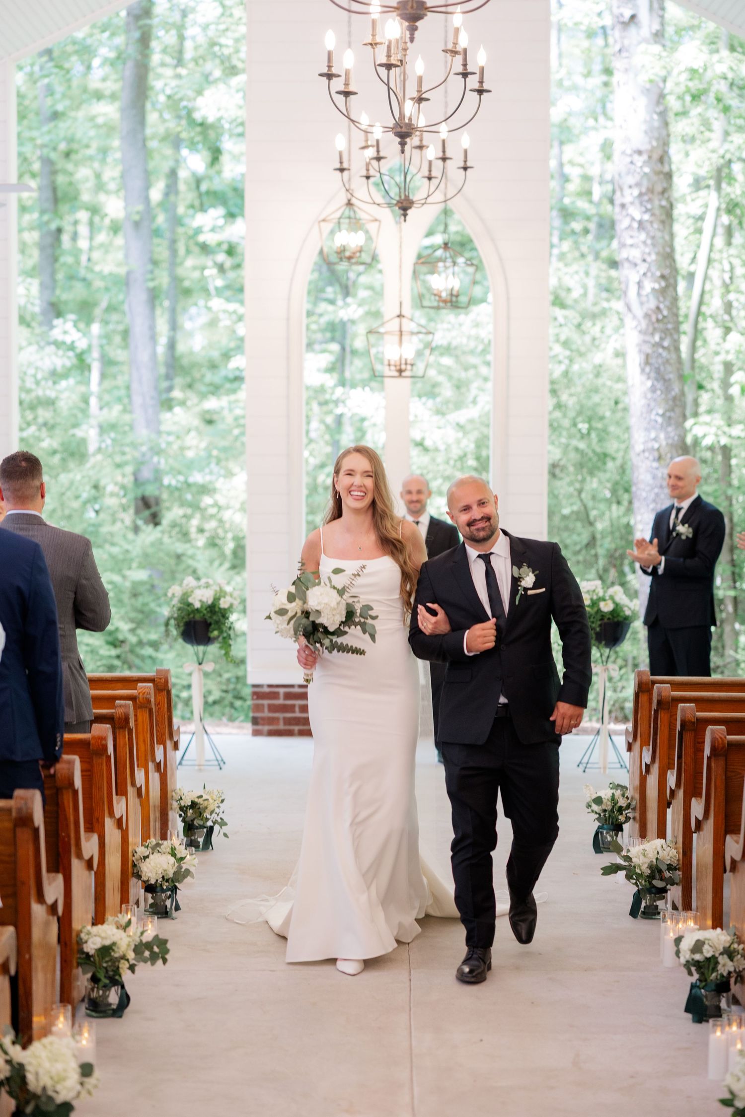 Elegant white wedding chapel interior with wooden pews and chandeliers surrounded by forest views.