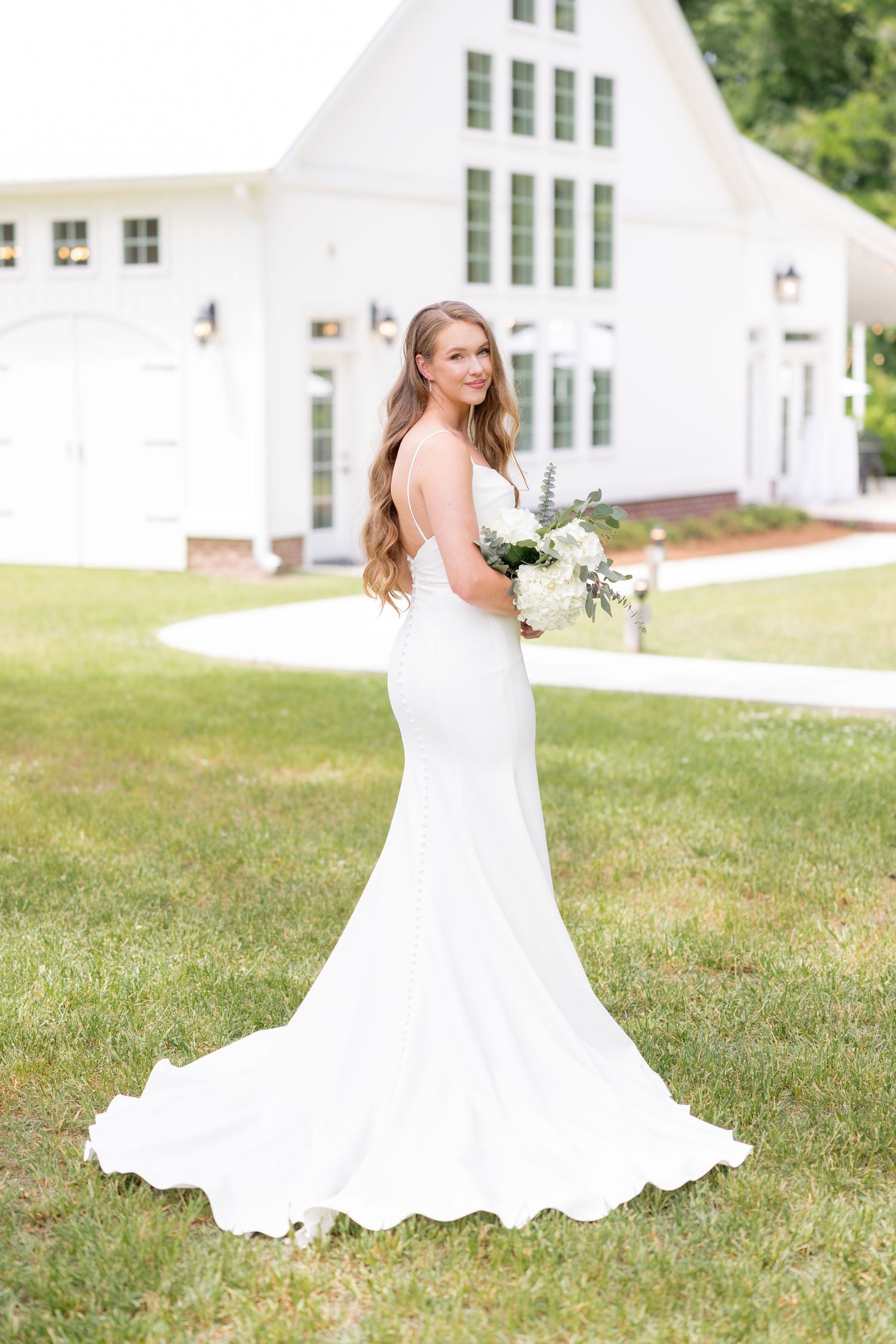 A bride in a flowing white wedding dress poses gracefully on a grassy lawn with a white chapel in the background.