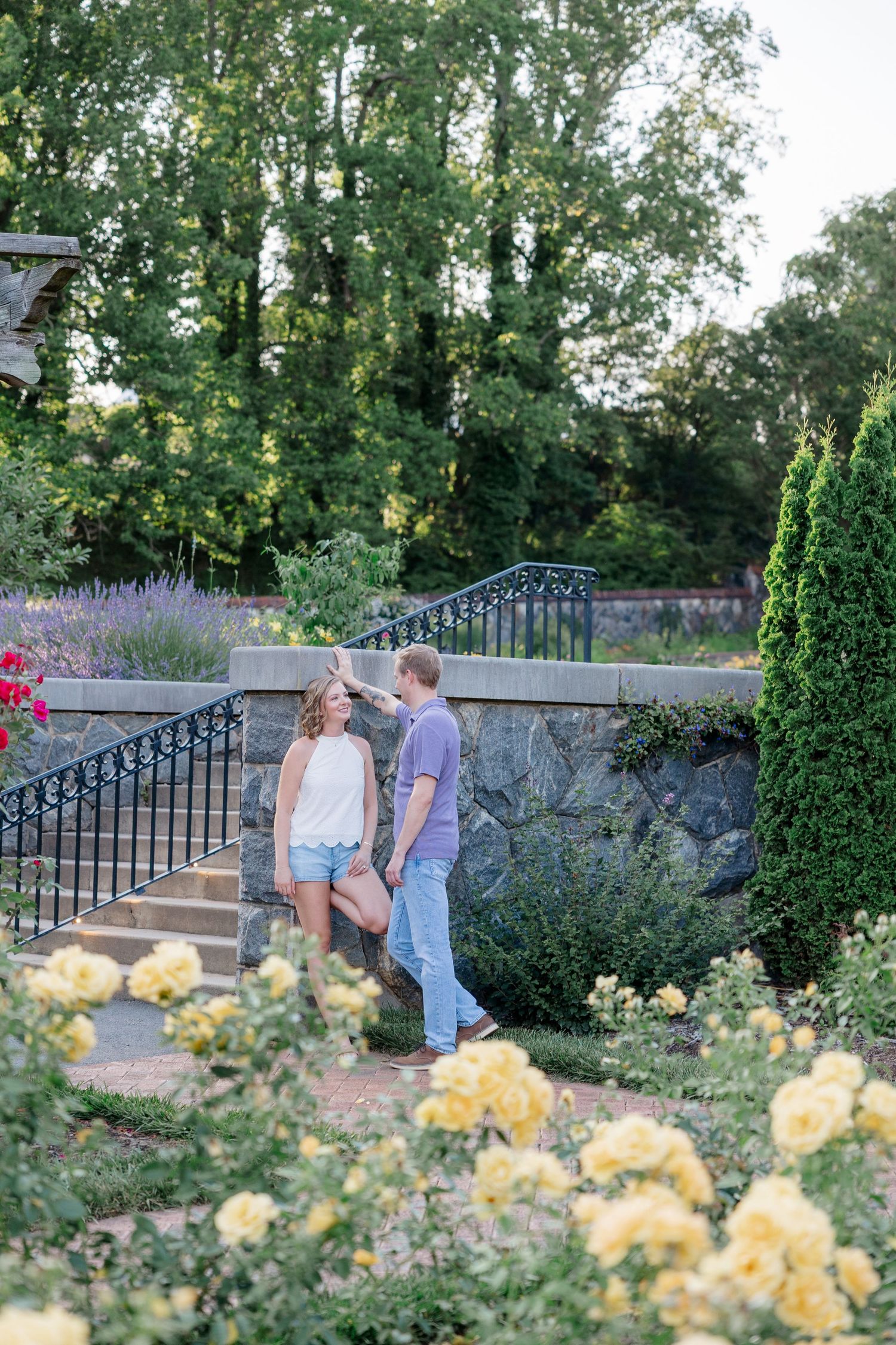 A couple shares a romantic moment in a garden with yellow roses and stone walls at sunset.